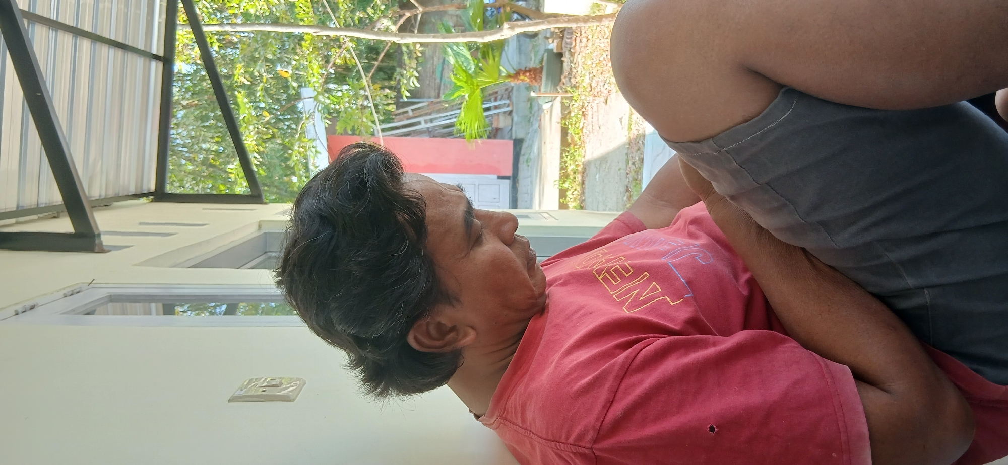 A side profile of a middle-aged Indonesian man is captured in an outdoor setting, possibly a patio or porch. He is seated, facing to his right and looking downwards, with his head tilted. The man has dark, wavy hair, styled with volume at the top and sides. He is wearing a faded red t-shirt with some lettering visible, appearing to be a light yellow and blue font. A small hole is present on the left side of his shirt, near the shoulder. He is also wearing grey shorts. His right arm is resting on his lap, while his left arm is obscured by his body.
The environment behind him suggests a residential area in Pejaten, Indonesia. A white wall and a window are visible to his left. To his right, the background shows a slightly blurred scene with lush green trees and foliage. A pink building with a white door and a ladder leaning against it can be seen in the distance. Sunlight illuminates the scene, casting shadows and highlighting the textures of the man's clothing and skin. The overall impression is a candid moment of relaxation or contemplation in a warm, tropical climate.
