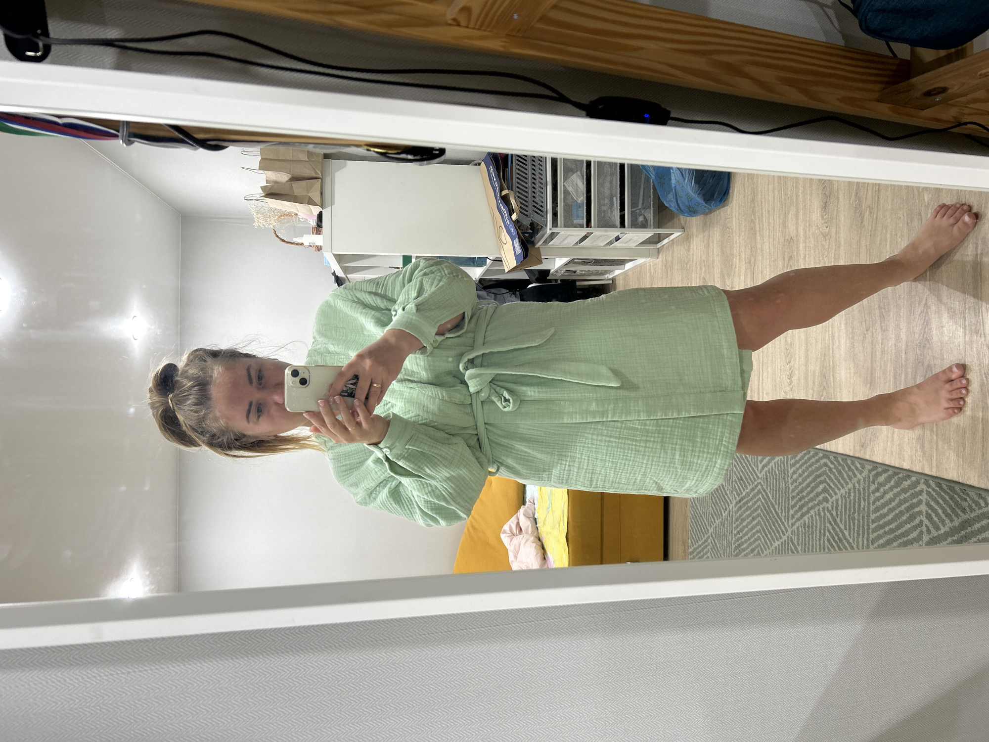 A young woman is taking a selfie in a mirror. She is wearing a light green robe and holding a white smartphone. The room appears to be a bedroom, with a bed, shelving unit, and rug visible. The location is not discernible from the image.
