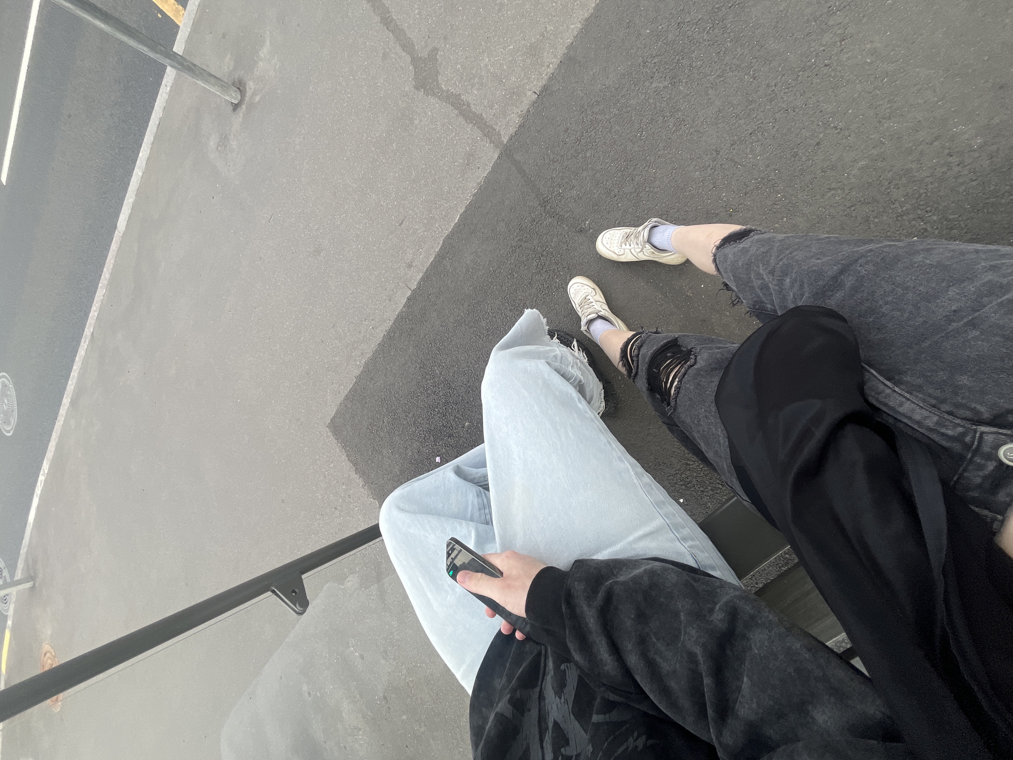 An overhead, slightly angled shot captures the lower torsos and legs of two individuals seated outdoors on a light grey concrete or asphalt surface in Saint Petersburg, Russia.
The person on the left wears a dark, possibly faded black, long-sleeved top and light blue jeans. Their legs are crossed, and they hold a black smartphone, which displays a green indicator, in their right hand. A black vertical metal pole, featuring a small bracket, is positioned to their left, suggesting a bench or railing structure.
The person on the right wears dark grey ripped jeans and white sneakers with light blue socks. A black garment or bag rests on their lap. Their feet are positioned on the dark grey asphalt ground.
The seating area (sidewalk) is separated by a distinct curb from a darker asphalt road. The road surface displays a white painted line and a partial yellow line further in the distance. Another lighter metallic pole is visible on the right side of the street, embedded in the ground. The general atmosphere suggests an urban outdoor setting.