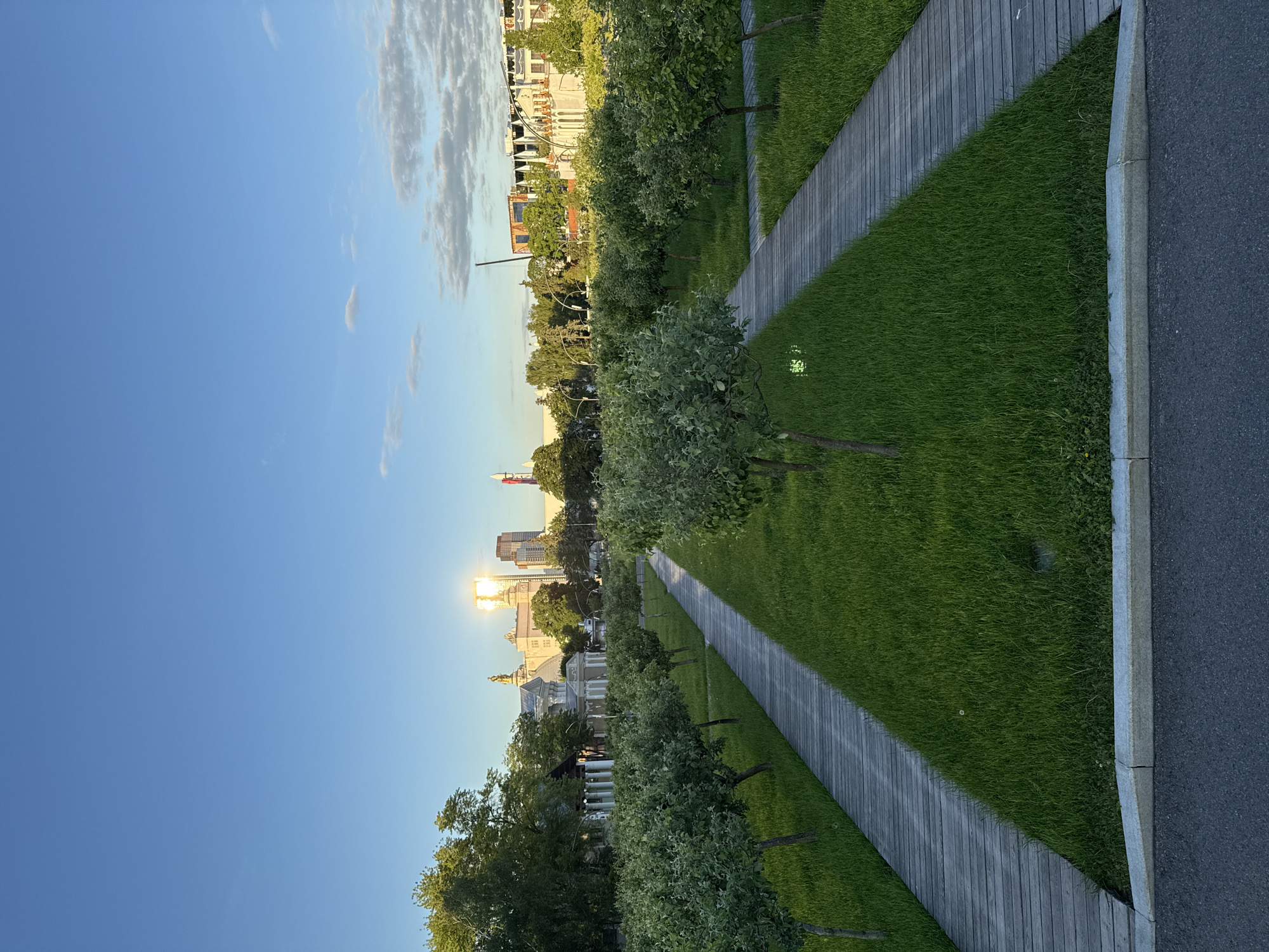 The image presents an expansive view of a well-maintained urban park leading towards a city skyline, captured during the golden hour. In the foreground, a diagonal wooden pathway stretches across a vibrant green lawn, flanked by neatly planted rows of small trees. To the right, the lawn meets an asphalt road, separated by a distinct concrete curb. Small, embedded ground lights are visible within the grass, suggesting evening illumination.
The midground features more of the park's lush greenery and trees, beyond which the cityscape unfolds. A diverse architectural landscape is visible, ranging from grand, ornate buildings with domes and classical elements (reminiscent of Moscow's historical structures) to modern, glass-clad skyscrapers. The setting sun, low in the sky, creates a brilliant glare off the reflective surface of a tall building, casting a golden hue across the scene.
The sky above is a clear, deep blue with scattered white, wispy clouds, indicative of fine weather. The strong, warm light suggests it is late afternoon or early evening. No people or specific activities are observable, allowing the viewer to focus on the interplay between nature and urban development. The combination of extensive green space, contemporary infrastructure, and distinctive historic architecture strongly points to a location within Moscow, Russia. There is no visible text in the image.