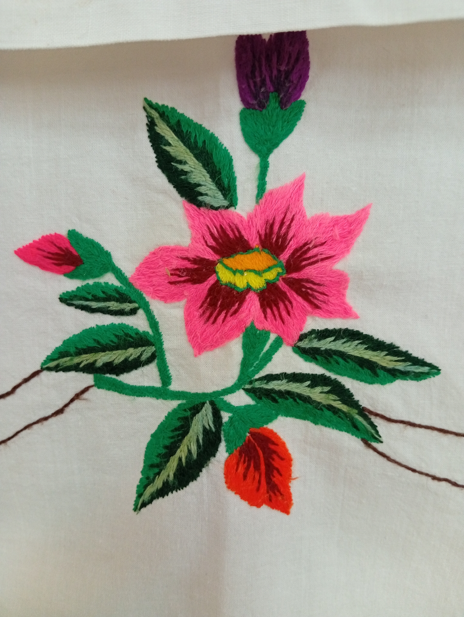 A close-up shot focuses on an embroidered floral design on white fabric. The embroidery features a large, vibrant pink flower with a yellow and green center. Surrounding the main flower are several green leaves, two pink buds, and one red bud. A purple flower bud is positioned above the pink flower. The embroidery is detailed with varied thread colors to create depth and texture. The white fabric appears to be linen or a similar natural material. The image is framed to emphasize the embroidery work, with a shadow cast from an unseen fold of fabric at the top. The location in Крыўлянскі сельскі Савет, Belarus is not depicted in the image.