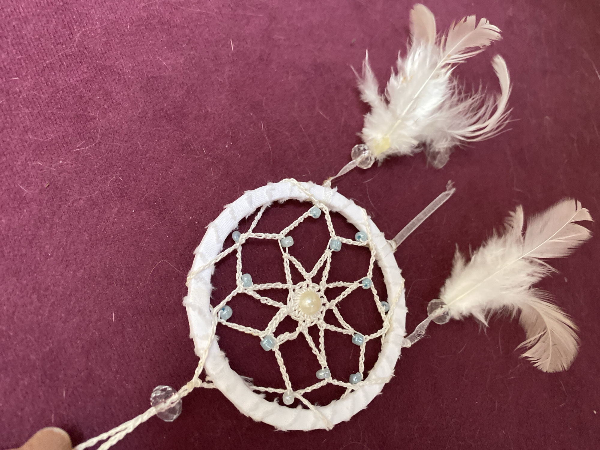 The image shows a close-up of a white dreamcatcher hanging against a dark red, textured background. The dreamcatcher has a circular hoop wrapped in white fabric, with a white web intricately crocheted inside. The web is adorned with small, light blue beads and a single, larger pearl in the center. Three delicate white feathers are attached to the bottom of the dreamcatcher, each suspended by a thin white string and a clear bead. A hand is visible in the upper left corner, holding the string that suspends the dreamcatcher. The background appears to be a soft fabric, possibly velvet or felt, with some fine white fibers scattered across its surface. The lighting is soft, highlighting the textures of the dreamcatcher and the background. There is no information about the location in Bílina, Czechia.