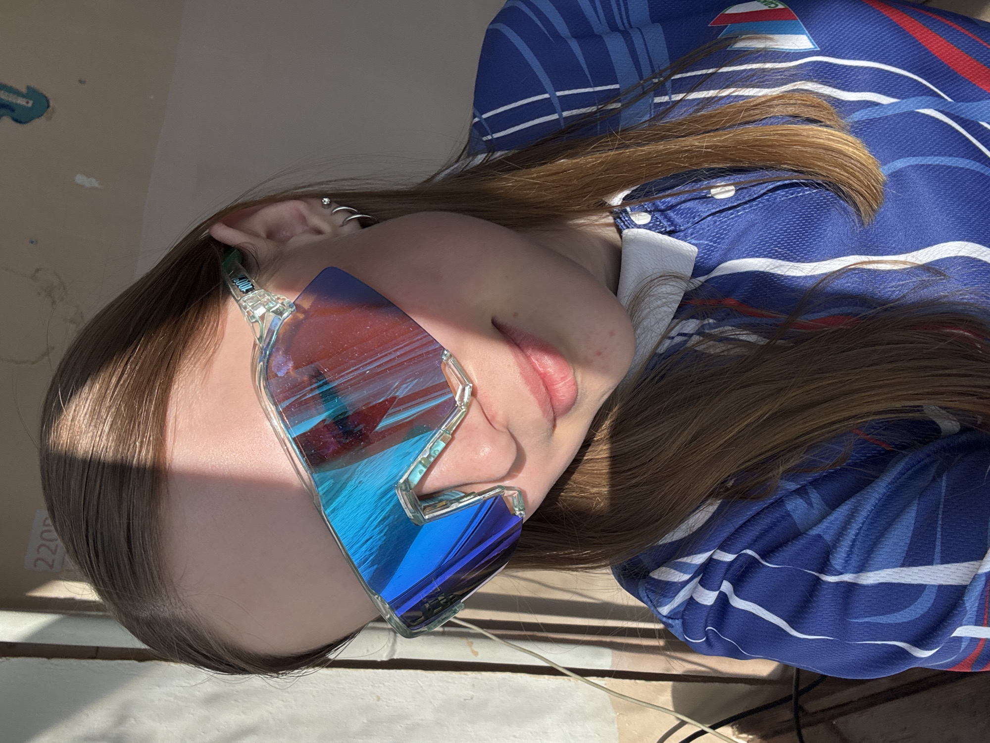 A young woman with long, light brown hair is wearing sunglasses with red and blue reflective lenses. She is wearing a blue polo shirt with white stripes and a white collar. She is positioned next to what appears to be a wall painted with a patch of light blue. The background is blurred, but a glimpse of an electrical cord is visible. The location is unknown and there is no evidence the image was taken in Berezniki, Russia.