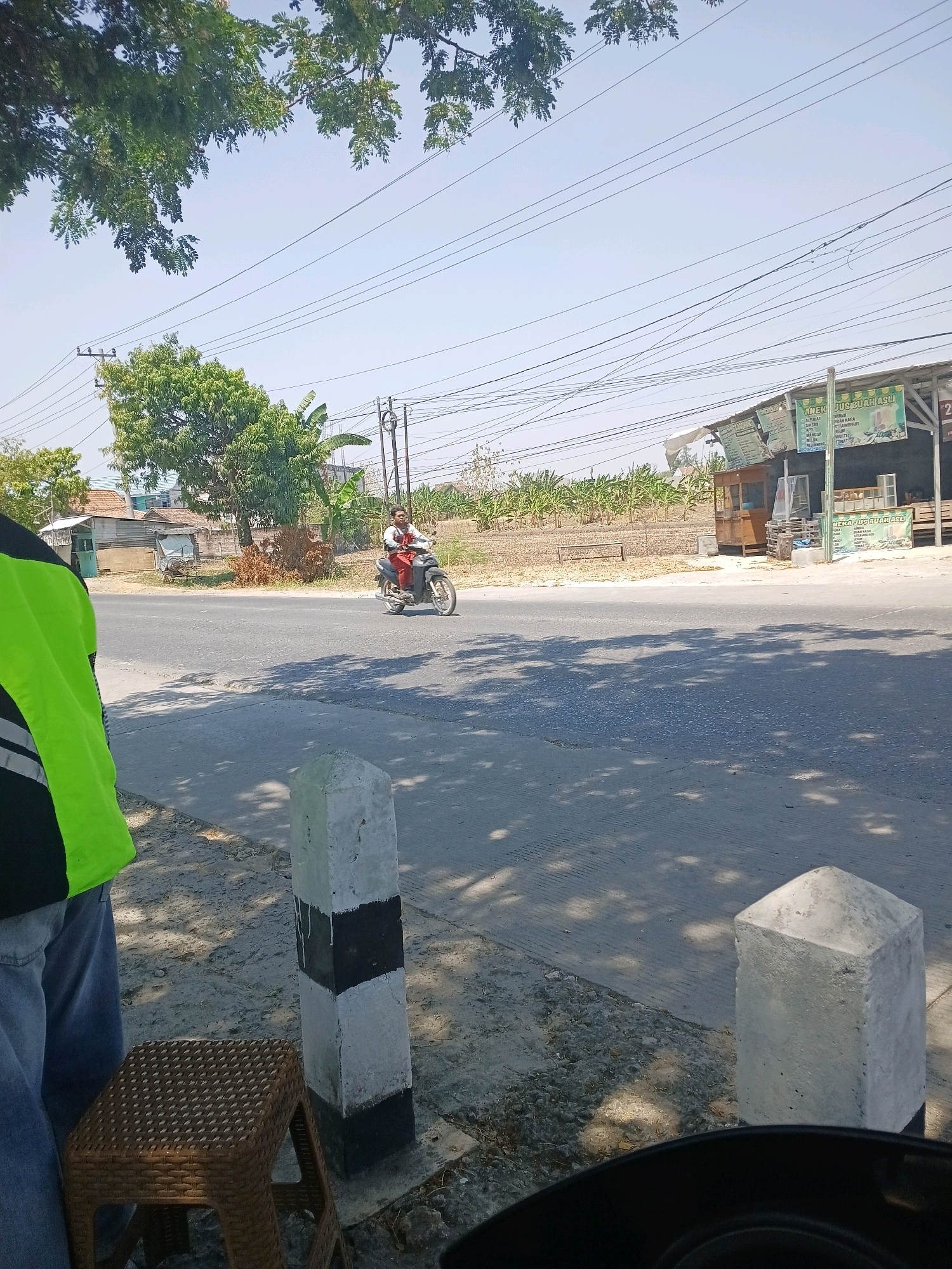 A person rides a motorcycle down a paved road. The person is wearing a red shirt and red pants and is looking forward. The motorcycle is dark colored. The road is lined with trees and there is a building to the right of the road. The building is a small shop with a sign that says "ANEK JUS BUAH ASLI". The sky is clear and blue. There are power lines crossing the sky. The scene is lit by bright sunlight.