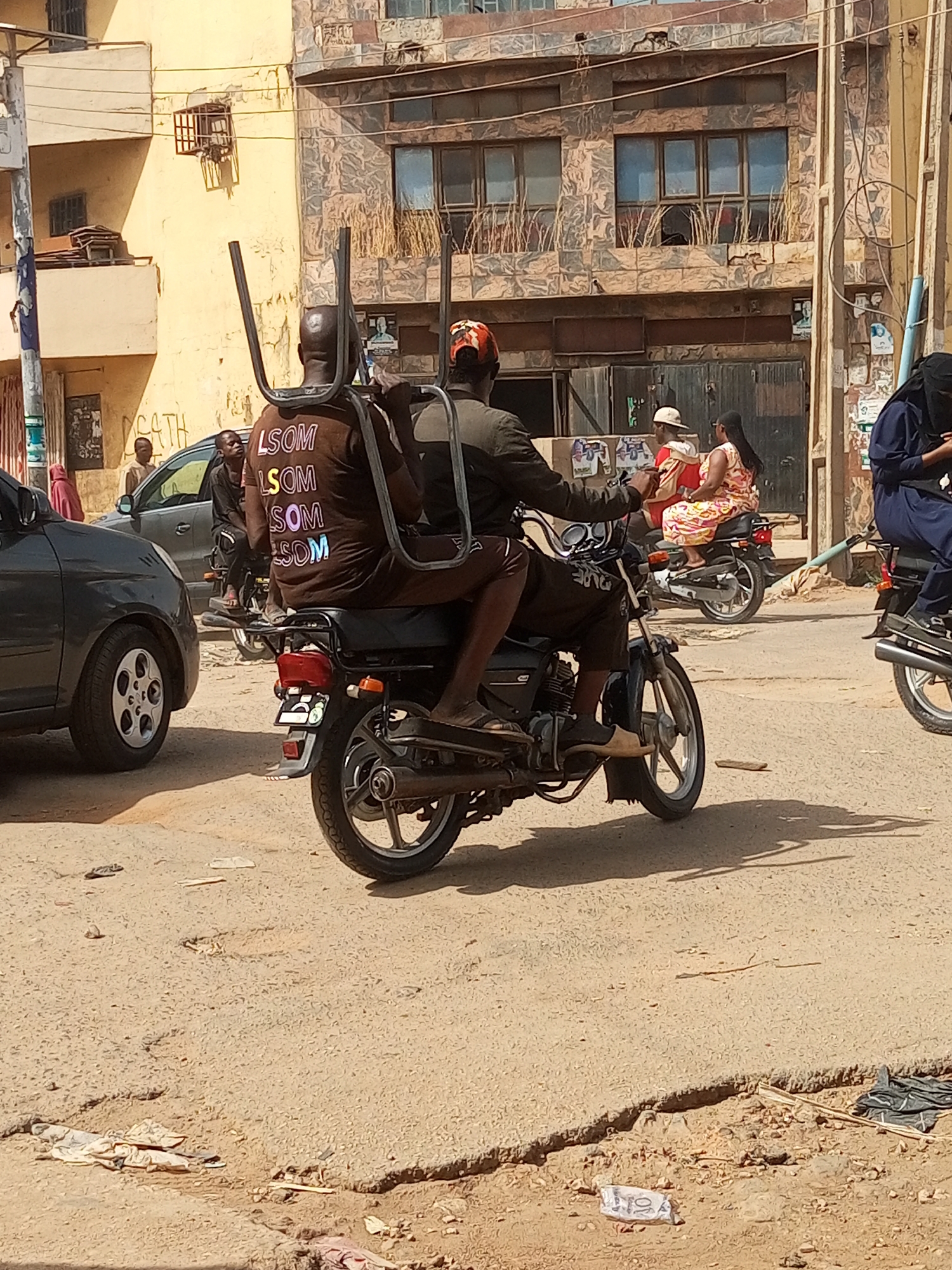 Location: Maiburiji, Nigeria.
Scene: A street scene with multiple people riding motorcycles. One motorcycle carries two chairs strapped to the back. The passenger wears a brown shirt with the letters "LSOM" repeated multiple times in different colors. Cars and pedestrians are also present. The road surface is cracked and littered with debris. The buildings in the background appear worn and multi-storied. Other motorcycles are also present in the scene. One rider wears a hijab.