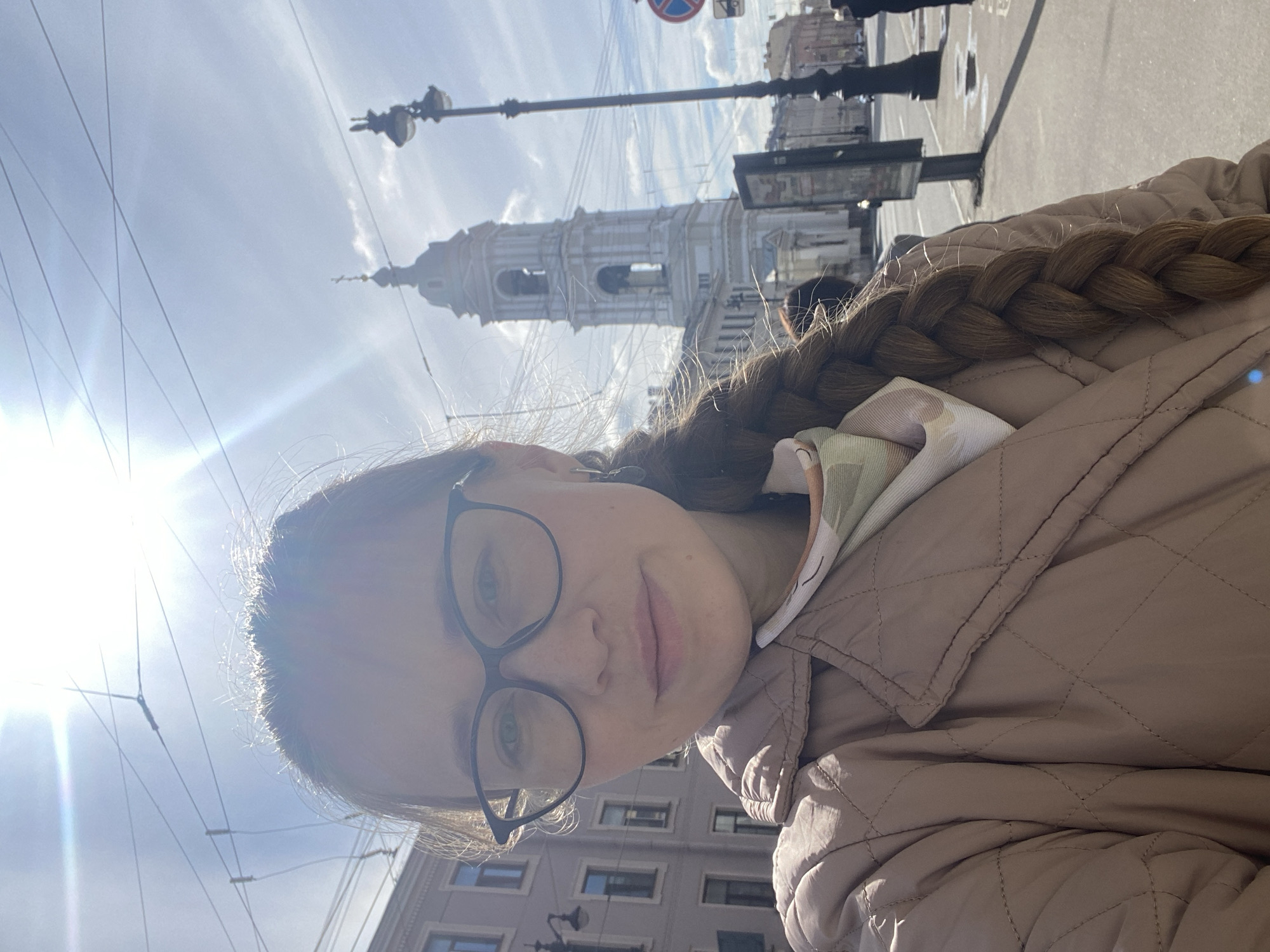 The image is a selfie of a young woman with a braided hairstyle, wearing glasses and a light-brown quilted jacket. She is standing outdoors in Saint Petersburg, Russia. In the background, there is a tall, white, multi-tiered baroque building with a spire, likely a church or cathedral. Streetlights and power lines are visible in the background. Buildings, including a structure with multiple windows, stand nearby the woman. The weather is sunny.