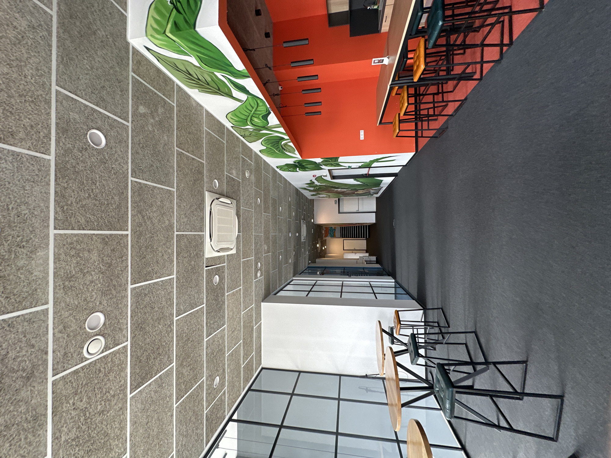 A slightly distorted, upward-angled interior shot depicts a modern commercial space. The floor is composed of large, rough-textured grey tiles, punctuated by evenly spaced circular recessed lights. An air conditioning unit is mounted on the wall in the center of the frame.
The ceiling features a bold design with vibrant tropical leaf murals. Adjacent to the mural is a bright orange wall with several horizontal lights, and a dark, built-in structure above a light-wood countertop and bar-style seating.
On the right, the flooring shifts to a dark grey, and seating arrangements are evident. A contrasting white section with large windows frames the lower portion of the image, allowing natural light to enter the space. Black metal frames define the window panes.
Given the image's modern design and use of bright colors and natural elements, it could be a restaurant, a modern office, or a co-working space. The location being in Varna, Bulgaria, cannot be verified from this image alone.