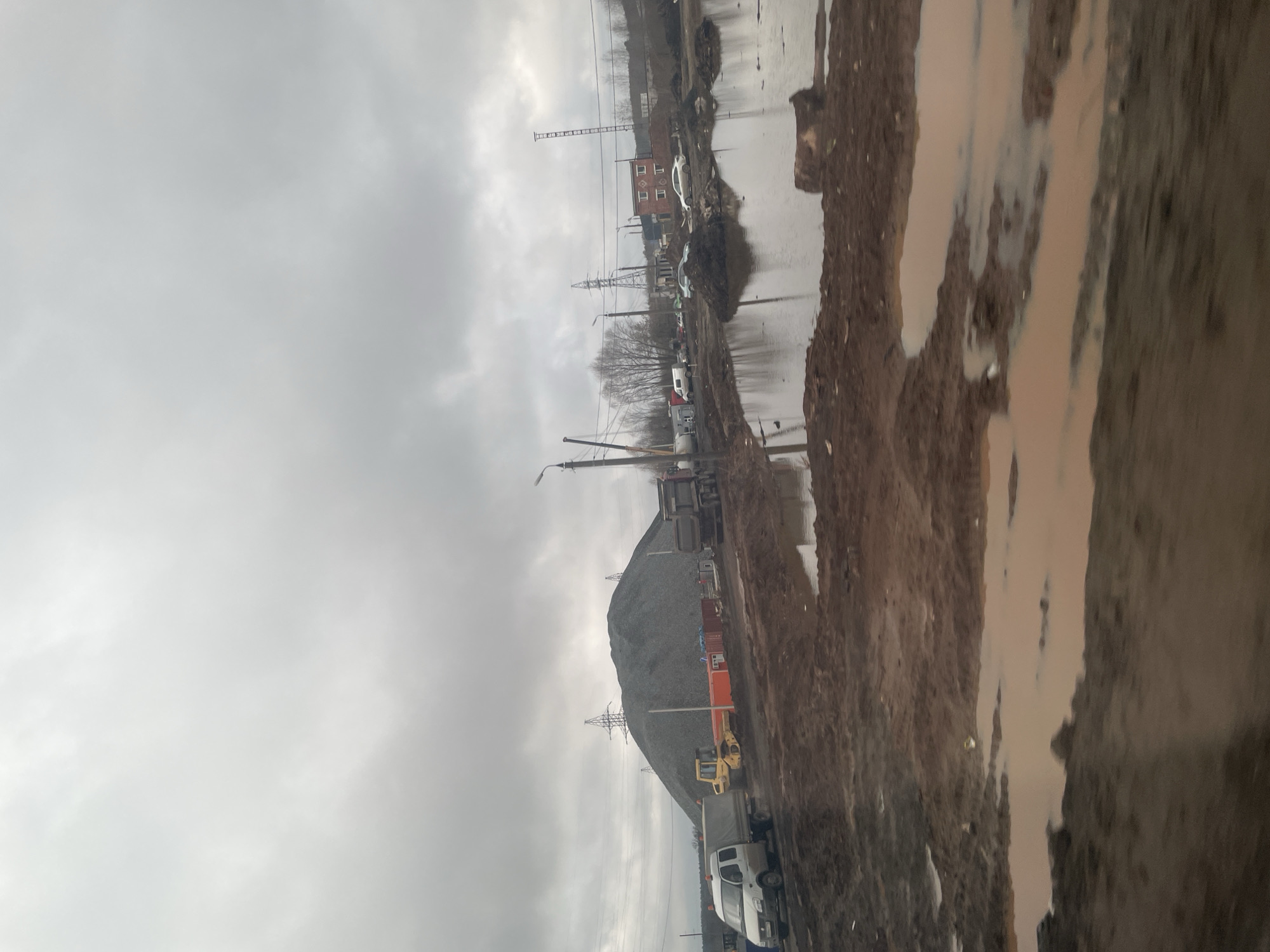 The image depicts a muddy industrial area in Kabanikha, Russia, under a cloudy sky. There's a large pile of gravel or construction material in the middle distance. Construction vehicles, including trucks and a bulldozer, are visible near the pile. In the background, buildings and power lines can be seen. The foreground is dominated by muddy terrain, with puddles reflecting the sky.