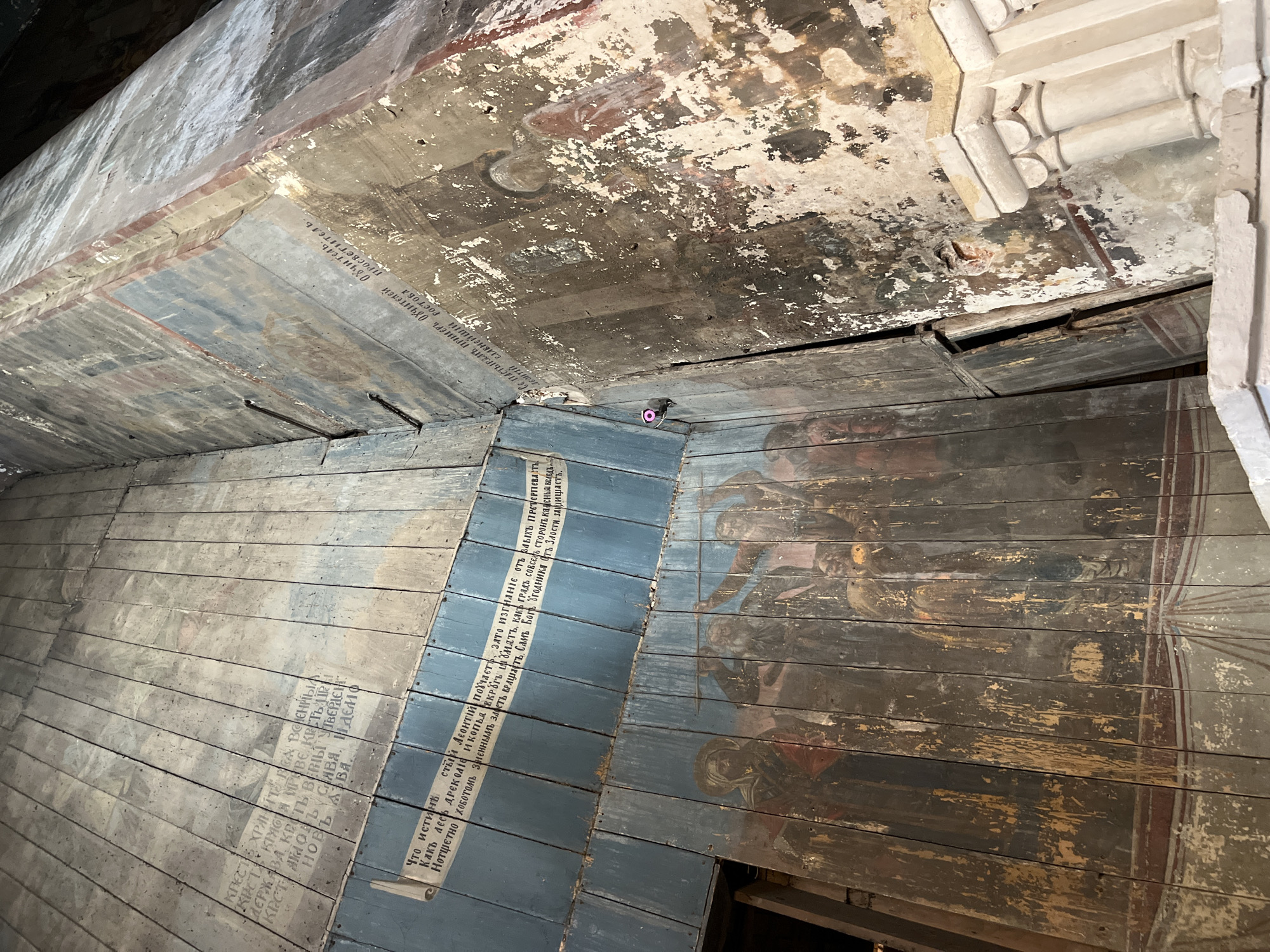The image shows the interior of a wooden structure with painted murals. The murals depict religious figures and text. The wood appears aged and worn, with some areas showing signs of decay and peeling paint. In the foreground, a group of figures is depicted in what appears to be a religious scene. To the right, the wall is heavily damaged, with paint flaking off to reveal the underlying material.
The text visible in the murals appears to be in Cyrillic script, suggesting a Russian origin. Based on the provided information, this scene is located in the city of Kladovitsy, Russia. The overall impression is one of historical significance and perhaps neglect, with the vibrant artwork contrasting with the decaying structure. A small pink camera or sensor is visible attached to the wooden paneling on the left side of the image.