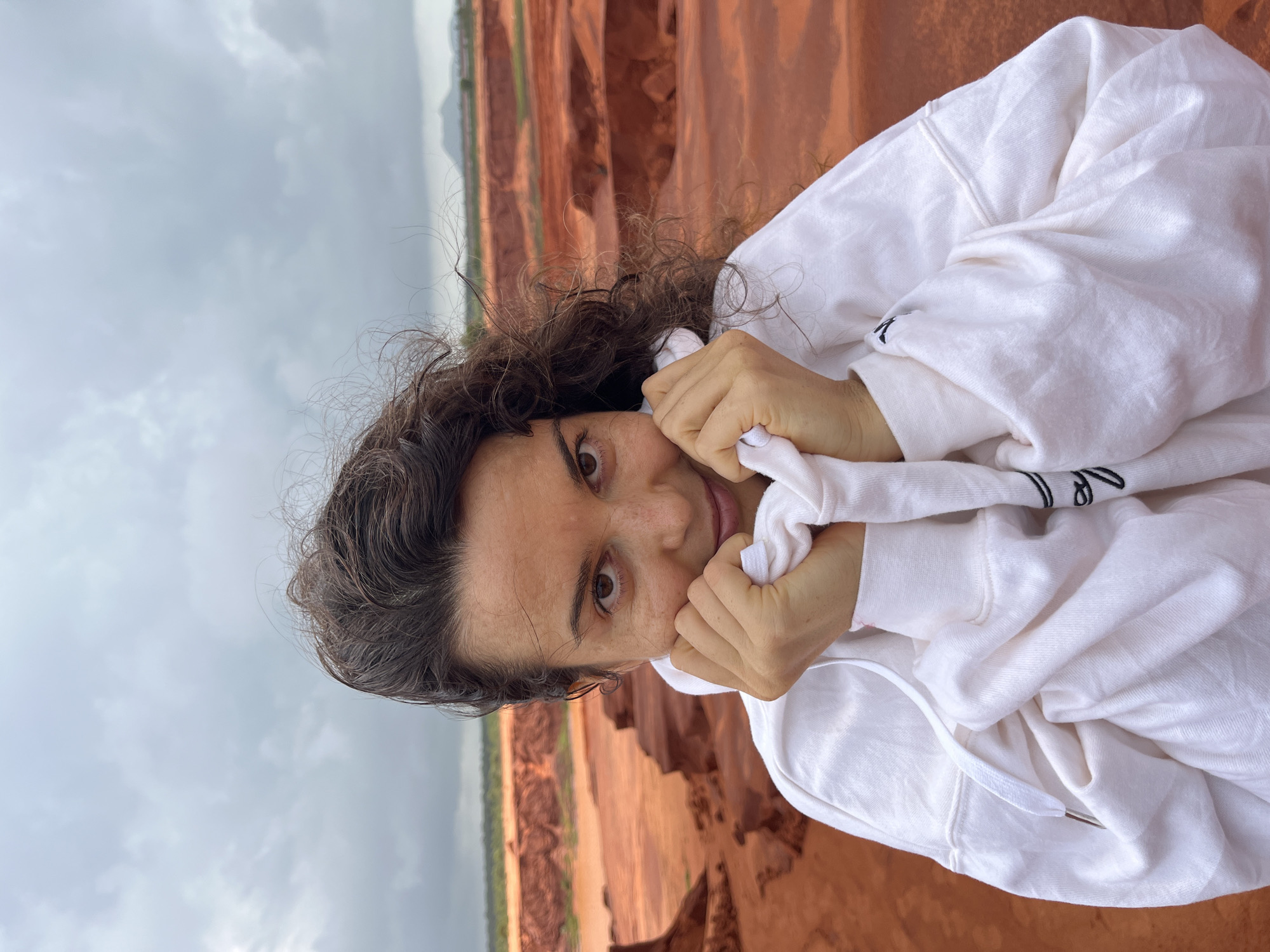 A woman with dark, curly hair pulled her white hoodie up to her chin, obscuring the lower half of her face. She had dark brown eyes and a smattering of freckles across her cheeks and nose. Her hands were clasped together, holding the hoodie in place. She was looking directly at the camera with a slight smile.
The background was a landscape of red rock formations, likely the Red Sand Dunes in Mui Ne, Vietnam. The sky was overcast, with grey clouds filling the upper portion of the frame. The contrast between the woman's bright white hoodie and the earthy red tones of the dunes was stark. The wind appeared to be blowing, as some of her hair was caught in the breeze.
