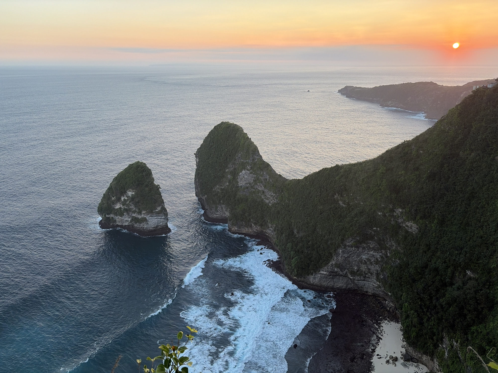 The image captures a panoramic view of the Kelingking Beach in Nusa Penida, Indonesia, during a sunset. The scene is dominated by the vast expanse of the ocean meeting a rugged coastline. Two prominent rock formations, covered in lush green vegetation, rise majestically from the turquoise waters on the left side of the frame. These formations are a defining feature of Kelingking Beach.
The coastline on the right is characterized by steep, verdant cliffs, also densely covered with foliage. The lower parts of these cliffs give way to a rocky shore and a small sandy beach, where waves are seen crashing with white foam. The water exhibits varying shades of blue and teal, with gentle ripples spreading across the surface.
The sky above is painted with soft hues of orange, pink, and yellow, indicating the setting sun. A small, bright sun disc is visible near the horizon on the right side of the image. The overall atmosphere is serene and natural, showcasing the dramatic beauty of this coastal landscape. There are no people or man-made objects, except for a few distant structures visible on the clifftop on the far right.