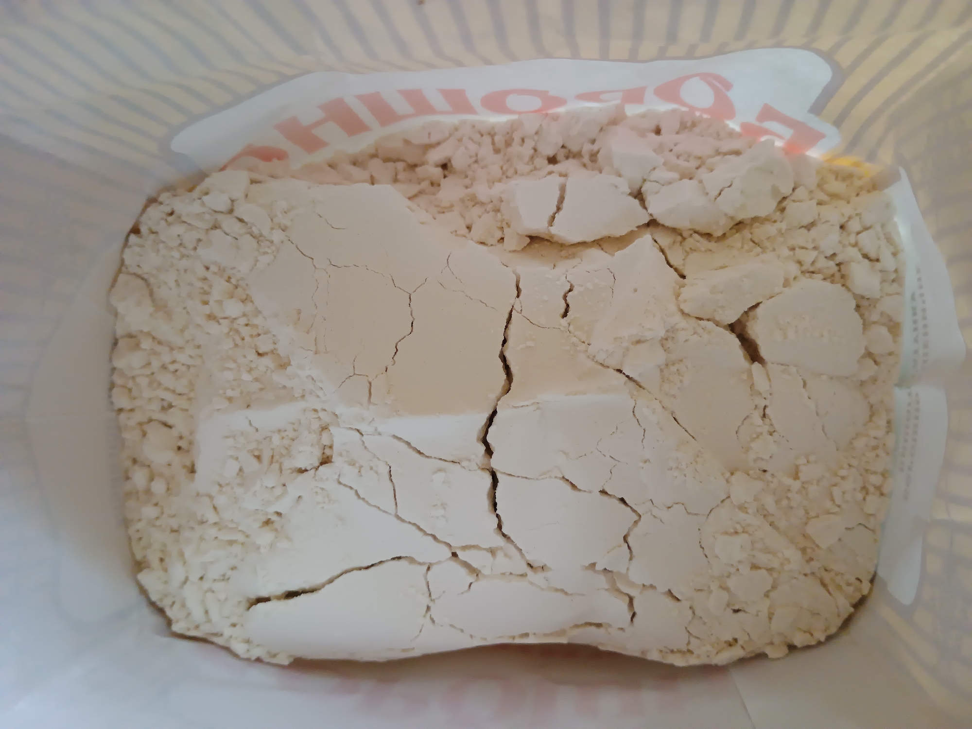 A close-up, top-down shot shows a pile of powdered food product, possibly flour, within a paper bag. The product is a light beige color and appears clumpy, with visible cracks and fissures running through it. Some smaller granular pieces are also present. The inside of the paper bag is visible, with white and yellow striped walls. A portion of the bag's printed label is visible on the right side, featuring red lettering and a partial pink design. The scene is devoid of any people or specific environmental details indicating a location in Zaporizhzhia, Ukraine. The lighting is bright, suggesting it was taken indoors or in daylight.