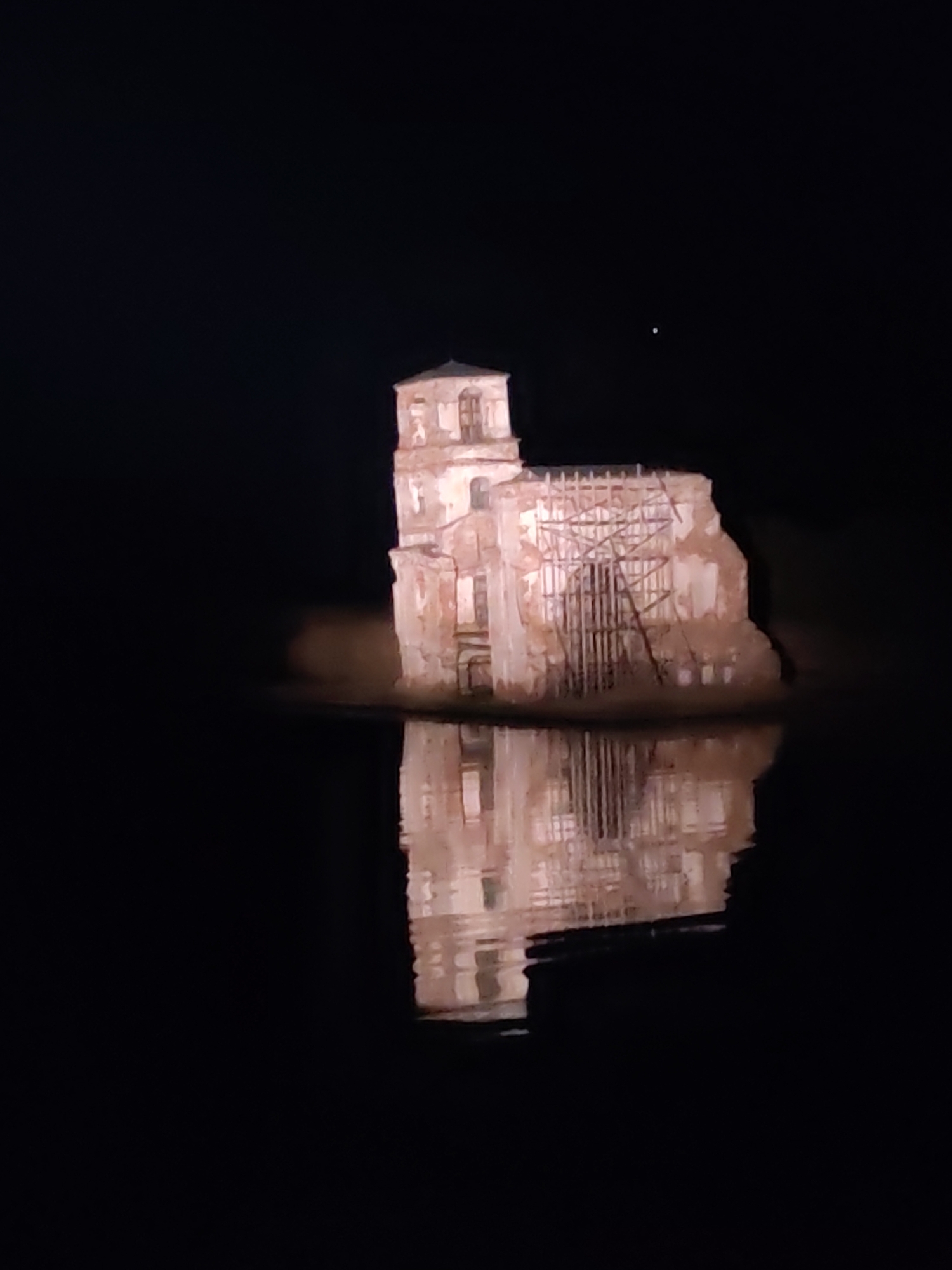 The image depicts the partially submerged ruins of the Church of the Nativity in Krokhino, Russia, at night. The building is illuminated and its reflection is visible in the surrounding water. Scaffolding supports one side of the structure. A single bright light, possibly a star or other celestial object, is visible in the otherwise dark sky.