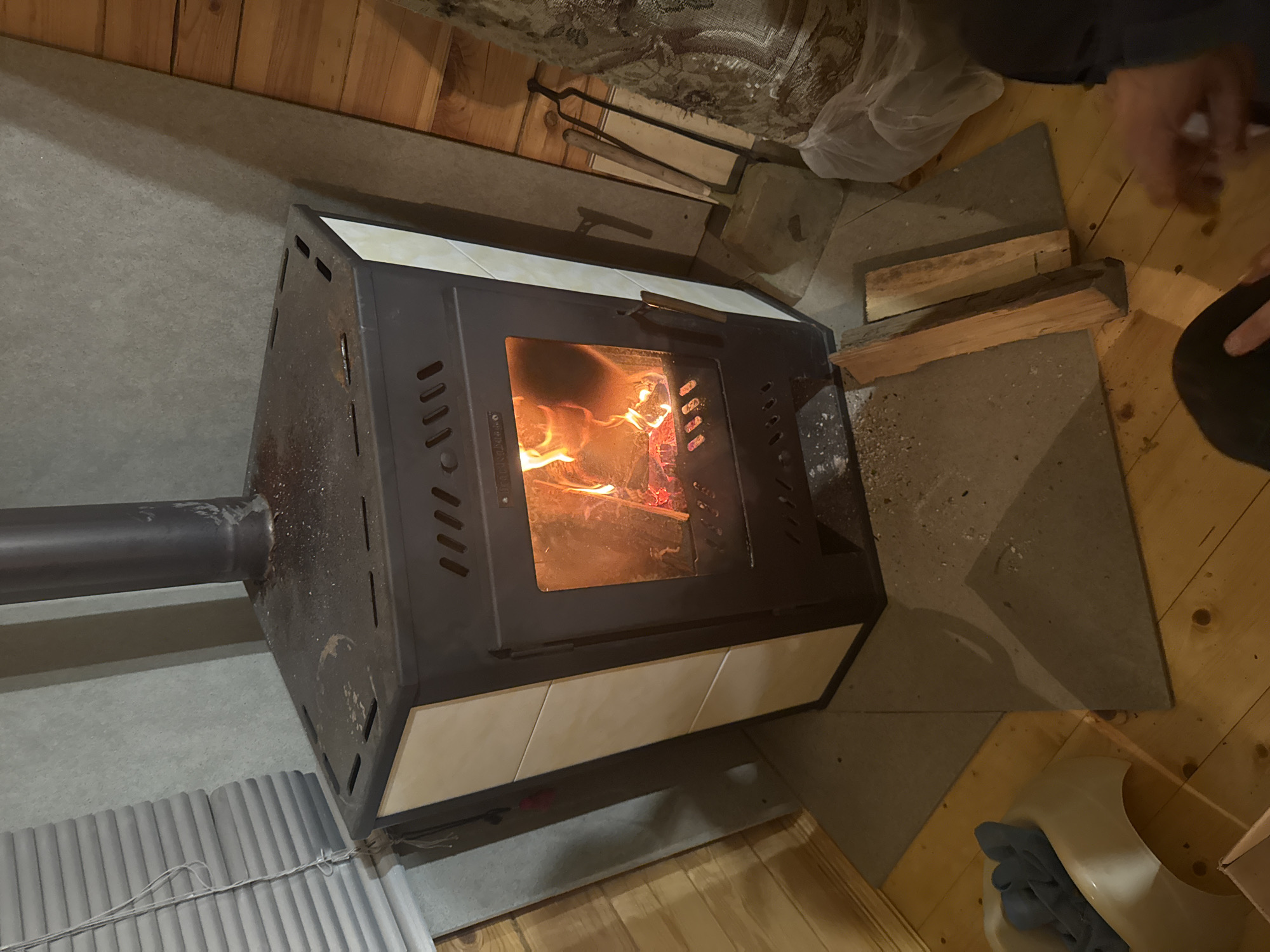The image depicts a wood-burning stove with a fire burning inside, located indoors on a tiled floor. The stove is black with cream-colored tiles around its base. A metal chimney extends from the stove to the left. Two pieces of wood are stacked to the right of the stove on the floor. A person's hand is visible in the upper-right corner. Other objects in the scene include corrugated metal sheets, a bucket, and a carpeted area, suggesting a domestic interior. There is no verifiable indication in the image that suggests the city of Mayak, Russia.