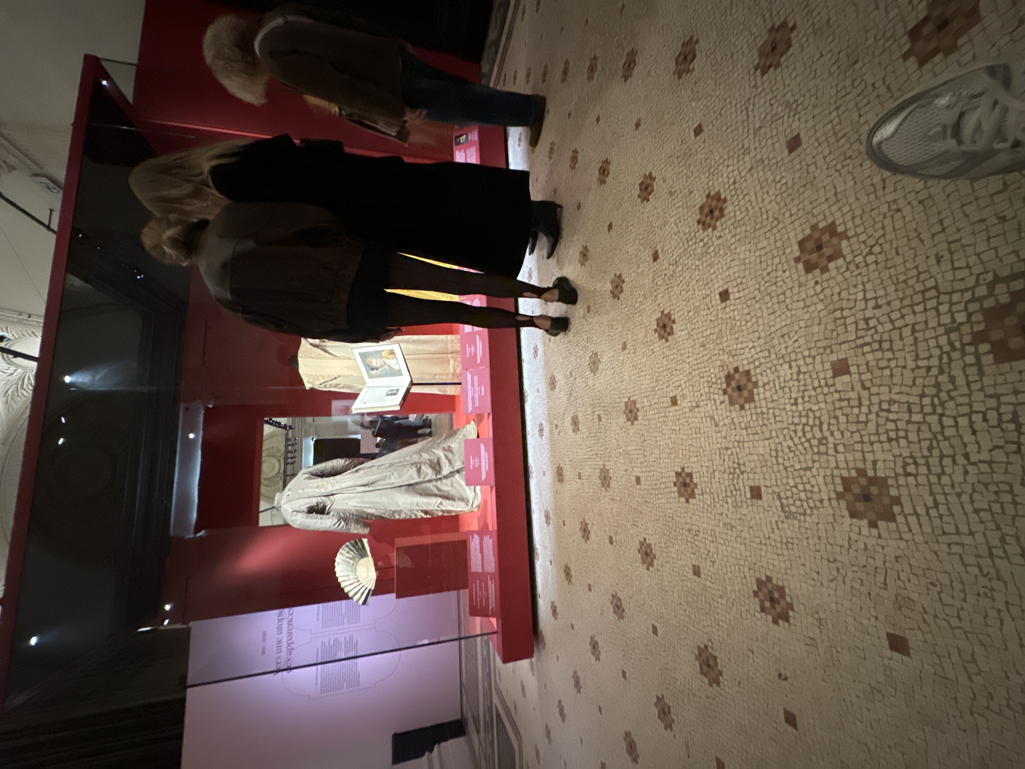 The image shows an interior space, likely a museum exhibit, located in Paris, France. Several individuals are observing artifacts displayed in a red-accented glass case. One person wears a brown jacket with fur trim, while another is clad in a black coat. A woman with exposed legs wears a gray jacket. The floor is adorned with a patterned mosaic. The museum showcases historical garments and related items, including a fan and displayed text. The scene conveys an atmosphere of cultural exploration and historical appreciation.