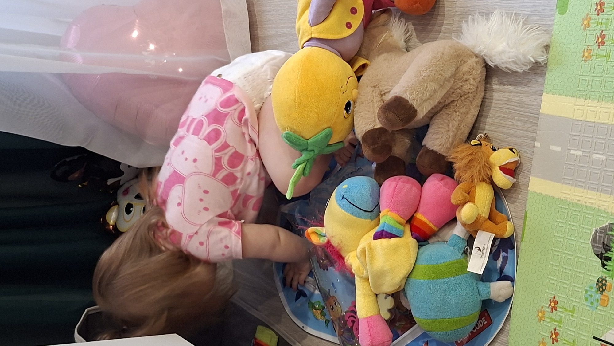 A young child wearing a pink and white bunny-patterned shirt is partially visible beneath a white fabric, possibly a sheet or curtain, with a pink balloon above it. The child is surrounded by a collection of stuffed animals, including a yellow flower-shaped toy, a beige horse, and a colorful plush creature, all situated on a blue circular play mat with underwater-themed illustrations. The play mat is positioned on a light gray floor, next to a green and yellow play mat with a road design. The setting appears to be indoors, possibly in a child's room. The location is Chaikovsky, Russia.