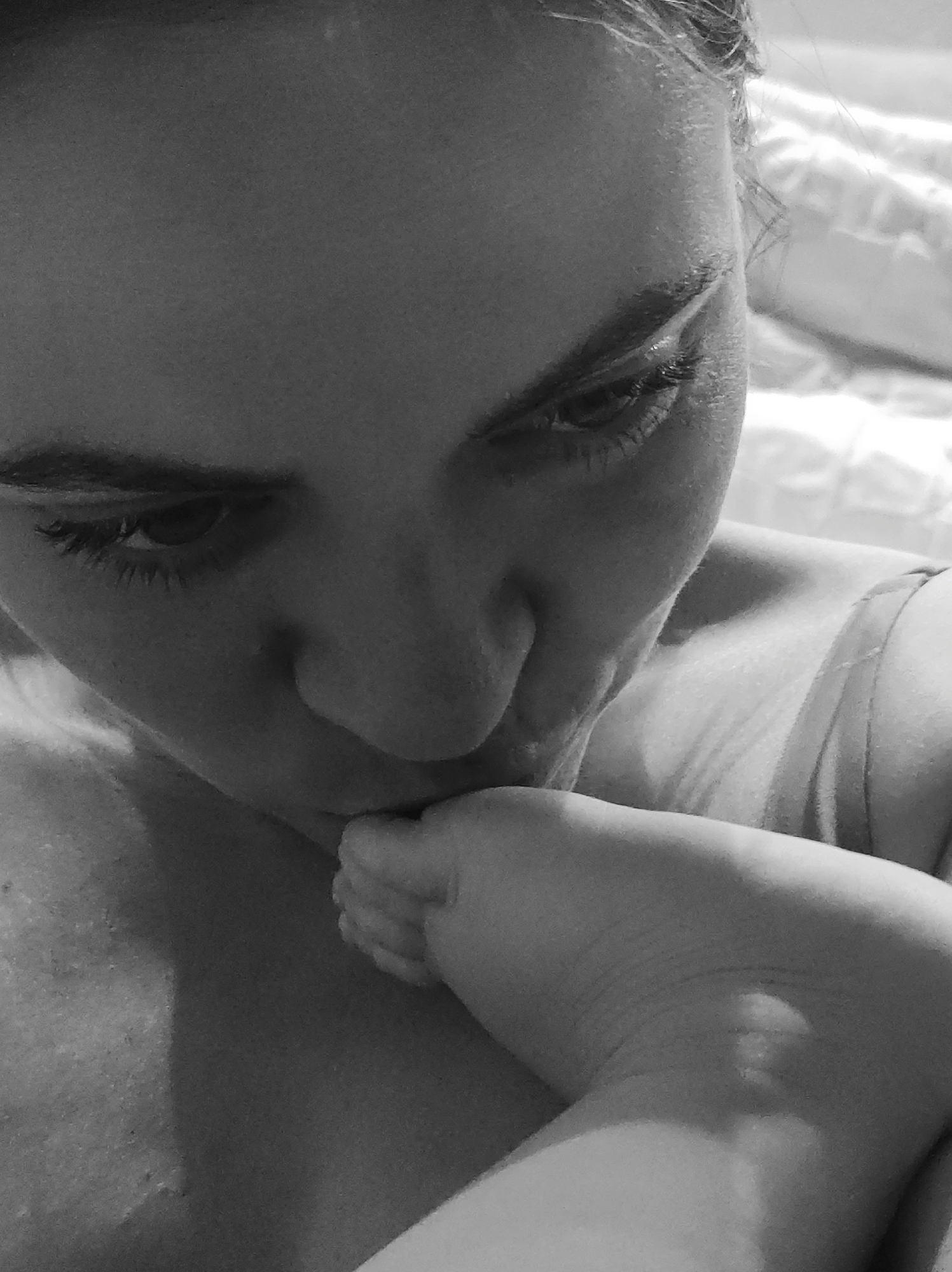 A grayscale, close-up image shows a woman’s face angled downward as she kisses a baby’s foot. The woman has long eyelashes and is wearing a tank top. The baby's foot is small and has several toes visible. The background includes what appears to be a white bed and bedding. It is not possible to confirm the location as Chaikovsky, Russia, from this image alone.