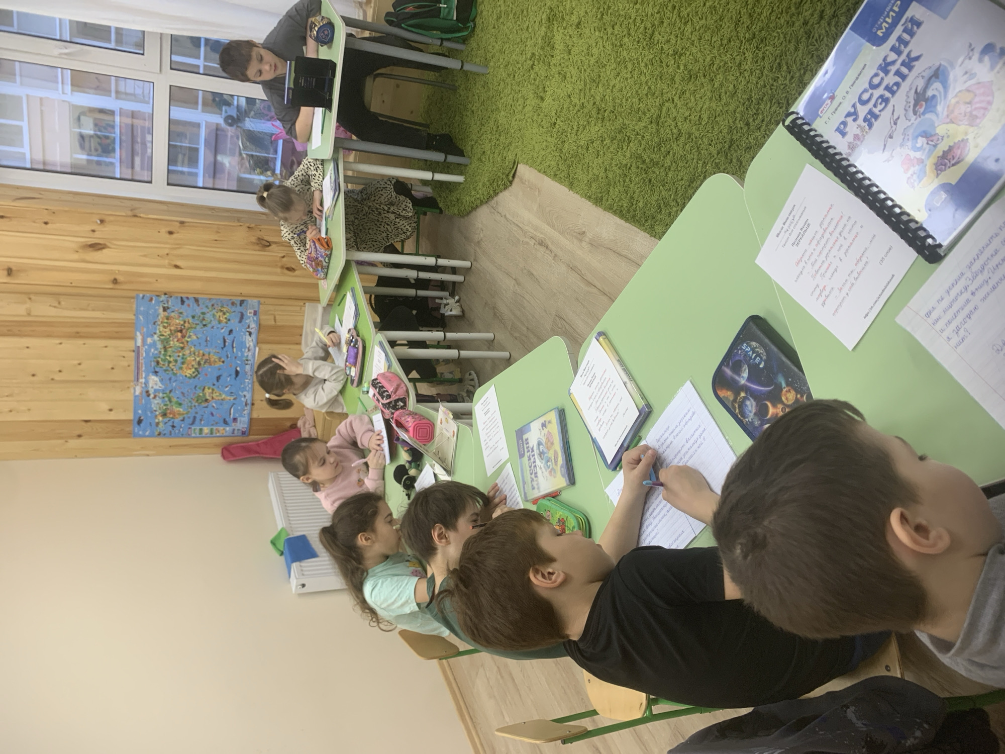 This media file depicts a vibrant classroom scene in Izhevsk, Russia, filled with primary school-aged children engaged in a learning activity. Approximately seven to eight children are visible, mostly seated at individual light green desks arranged in rows. The setting features light wooden wall paneling, a large window allowing natural light into the room, and a floor partially covered with a shaggy green carpet and partially with light wooden planks.
The children are actively involved in their studies, primarily writing in notebooks and referencing textbooks. In the foreground, two boys are seated, one diligently writing with a pen, while the other looks towards the front. Further down the desks, other students are focused on their work, including a girl in a leopard print top. At the far end of the room, a boy stands by a desk.
Notable objects include a colorful world map poster displayed on the wooden wall, various textbooks and notebooks on the desks, and colorful pencil cases. A prominent textbook on a desk in the foreground is titled "РУССКИЙ ЯЗЫК" (Russian Language) with "ОТКРЫВАЙ МИР" (Discover the World) visible on its cover, along with the authors "Г.Г. Граник, О. В. Гвинджилия." Several papers are visible on desks; one contains a printed Russian text titled "ЗЕРКАЛЬЦЕ" (Mirror) by Leonid Yakhnin, identified as "Текст для списывания" (Text for copying). Other papers display handwritten exercises in Russian, such as "нашла зеркальце" (found a mirror) and phrases like "не успела закрепить по" (didn't manage to fix by). The time of day appears to be daytime, given the natural light from the window. The children seem concentrated on their tasks, creating an atmosphere of focused learning.