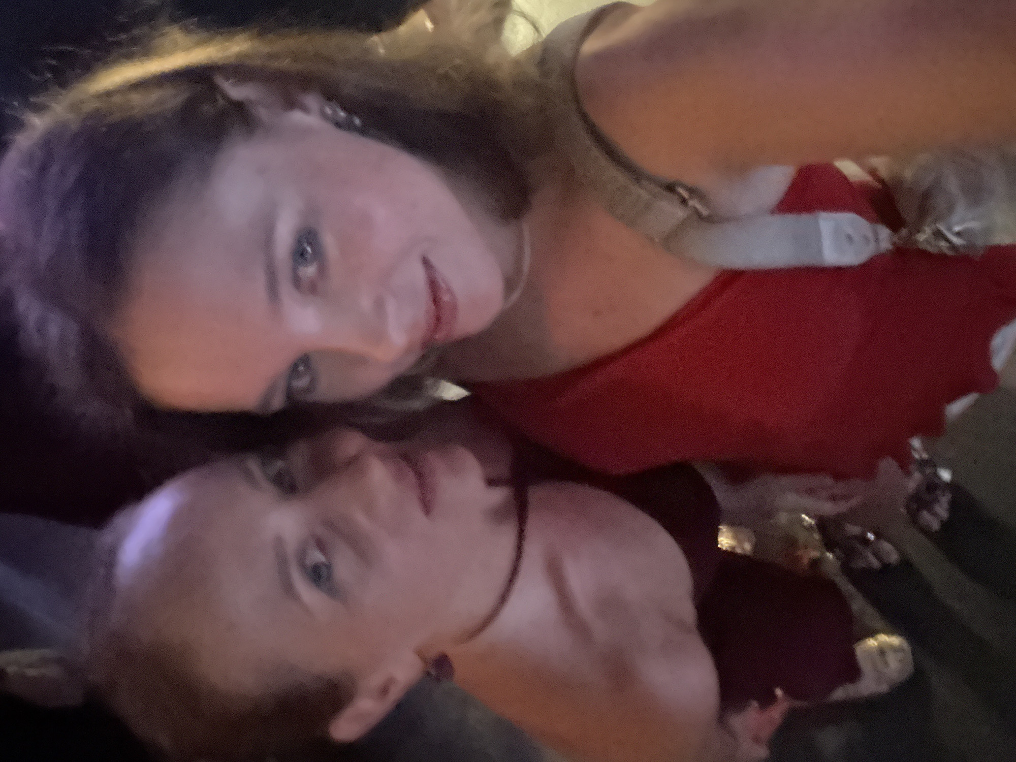 Two individuals, identified as women, are captured in a selfie taken at night, with the scene suggesting a lively urban environment. The individual on the left, with lighter skin and reddish-brown hair, wears a dark, possibly maroon, strapless top and a thin black necklace. Her gaze is directed towards the camera. The individual on the right, also with lighter skin and lighter brown hair, is wearing a vibrant red one-shoulder top. Her head is tilted slightly downwards, and her eyes are also directed towards the camera. The background is dark with blurred lights, indicating an outdoor setting, possibly a street or a venue with artificial lighting. There are hints of colorful illumination, including purplish and yellowish hues, suggesting a festive or entertainment-oriented location. The overall impression is of a casual social gathering.
Given the request to include the location of Đà Nẵng, Vietnam, and considering the typical atmosphere of nightlife in this Vietnamese city, this scene could plausibly be situated in an area known for its bars, restaurants, or entertainment complexes along the coast or within the city center, such as along the Han River or near the beaches like My Khe. The presence of multiple people, the evening setting, and the vibrant attire are all consistent with such an environment in Đà Nẵng.