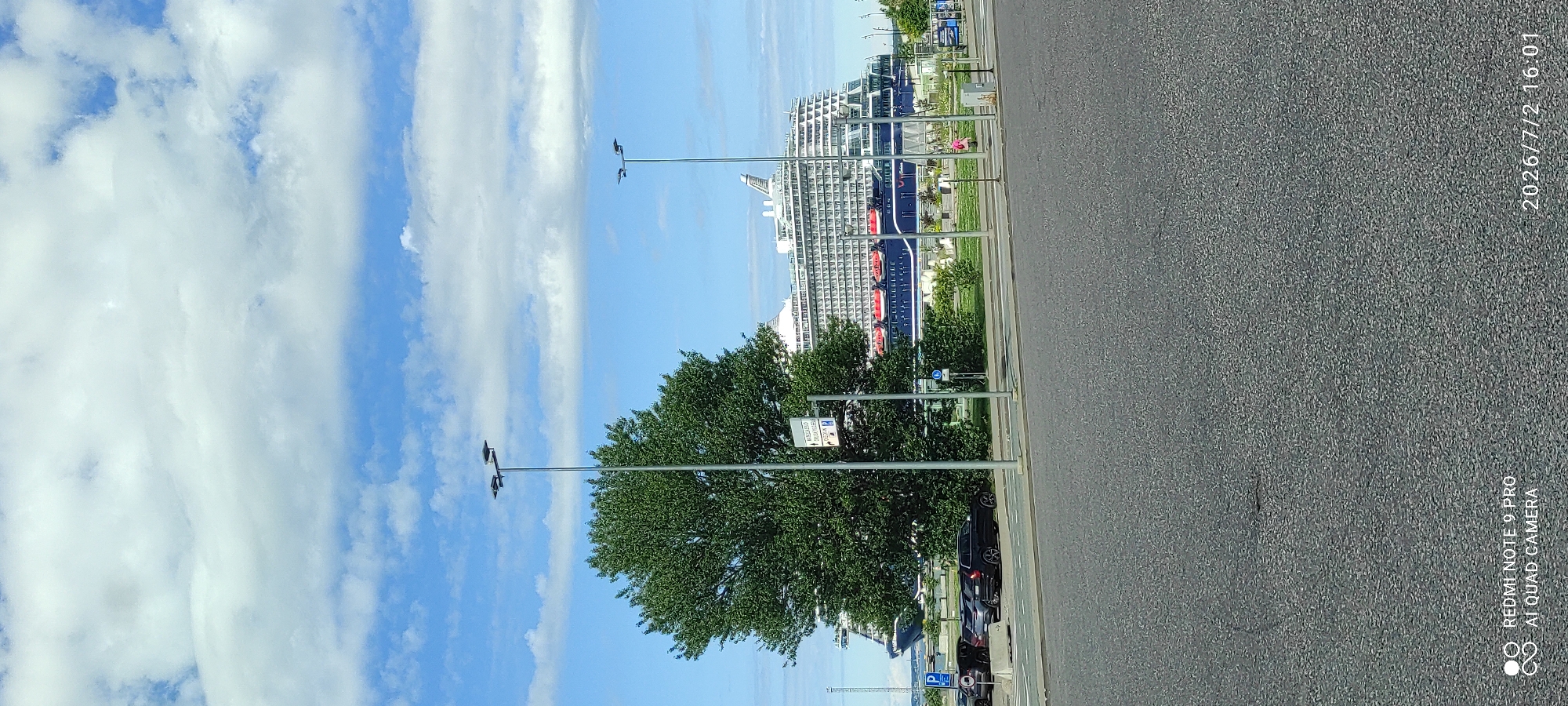 This media file shows a large cruise ship docked at a port. The ship is predominantly blue with white decks and red accents. It is a sunny day with a blue sky and scattered white clouds. The foreground features a paved road and a lush green tree. Several streetlights are visible, as are a few parked cars. The scene appears to be a public area with walkways and some signage. There is no discernible activity or event taking place, other than the ship being docked.
Notable details include the clear weather, suggesting it's daytime, likely afternoon given the timestamp "2026/7/2 16:01" in the corner of the image. The timestamp also indicates the date. The presence of the cruise ship and port facilities suggests an urban or coastal location. The overall mood is calm and serene.
Visible text includes the timestamp "2026/7/2 16:01" in the upper right corner. In the bottom right corner, there is a watermark with text "REDMI NOTE 9 PRO AI QUAD CAMERA". There are also some unreadable signs and labels on poles and possibly on the ship itself.