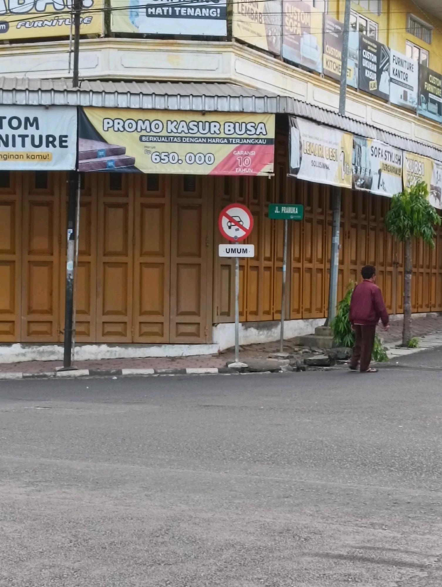 The image shows a street scene in Purworejo, Indonesia, with a row of shops lining the right side of the frame. The shops have large wooden doors and awnings displaying various advertisements. One banner prominently features "PROMO KASUR BUSA" (Foam Mattress Promo) with a price of "650.000" and a "GARANSI 10 TAHUN" (10 Year Warranty). Another banner to the left reads "TOM FURNITURE". Above the awnings, smaller banners advertise "CUSTOM SOFA", "CUSTOM KITCHEN", "FURNITURE KANTOR" (Office Furniture), and "CUSTOM UKURAN KASUR" (Custom Mattress Size). A street sign with "JL. PRAMUKA" is visible. A traffic sign indicating no cars is also present. A man in a maroon jacket and brown pants is walking away from the camera on the sidewalk, and a small tree is located next to the building. The street is paved, and there are some minor cracks and debris on the sidewalk.