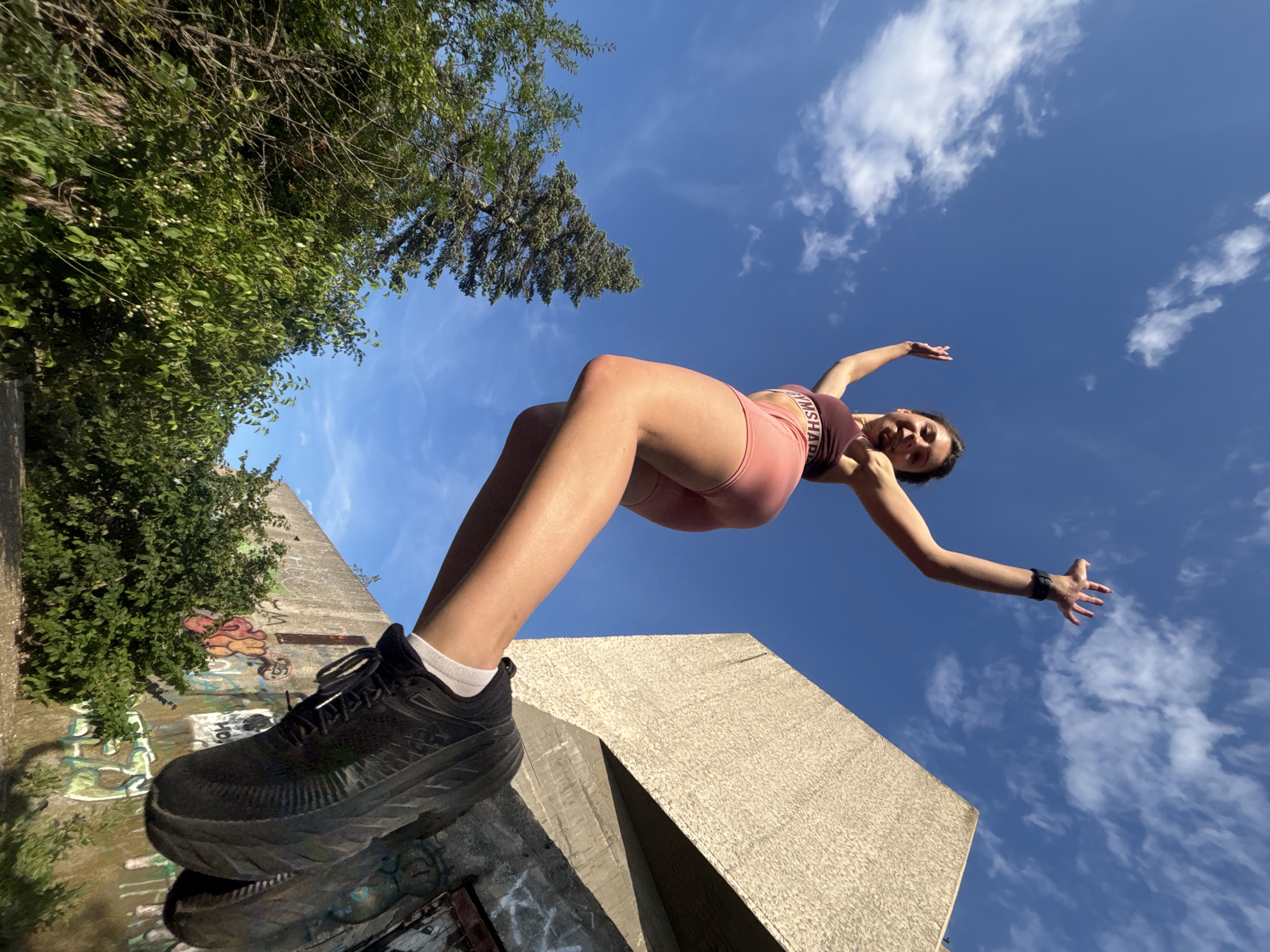 A young woman in athletic attire, a maroon sports bra and pink shorts, is captured mid-jump against a bright blue sky with scattered white clouds. She is positioned in the foreground, her body angled upwards and to the right, with her left leg extended forward and her right leg bent. Her arms are raised, with her left hand open and her right hand partially visible. She is wearing black athletic shoes with white socks.
In the background, a large, brutalist concrete structure dominates the upper right of the frame. The concrete appears weathered and is partially covered in graffiti. To the left of the woman, lush green trees and foliage are visible, with a tall evergreen tree standing out.
The perspective is low-angle, looking up at the woman from a position near the ground. The scene suggests an outdoor urban or semi-urban environment, possibly a park or an abandoned area. The image is sharp and well-lit, indicating daylight conditions. The overall impression is one of freedom and athleticism. The image was taken in the city of Varna, Bulgaria, likely near an architectural remnant.