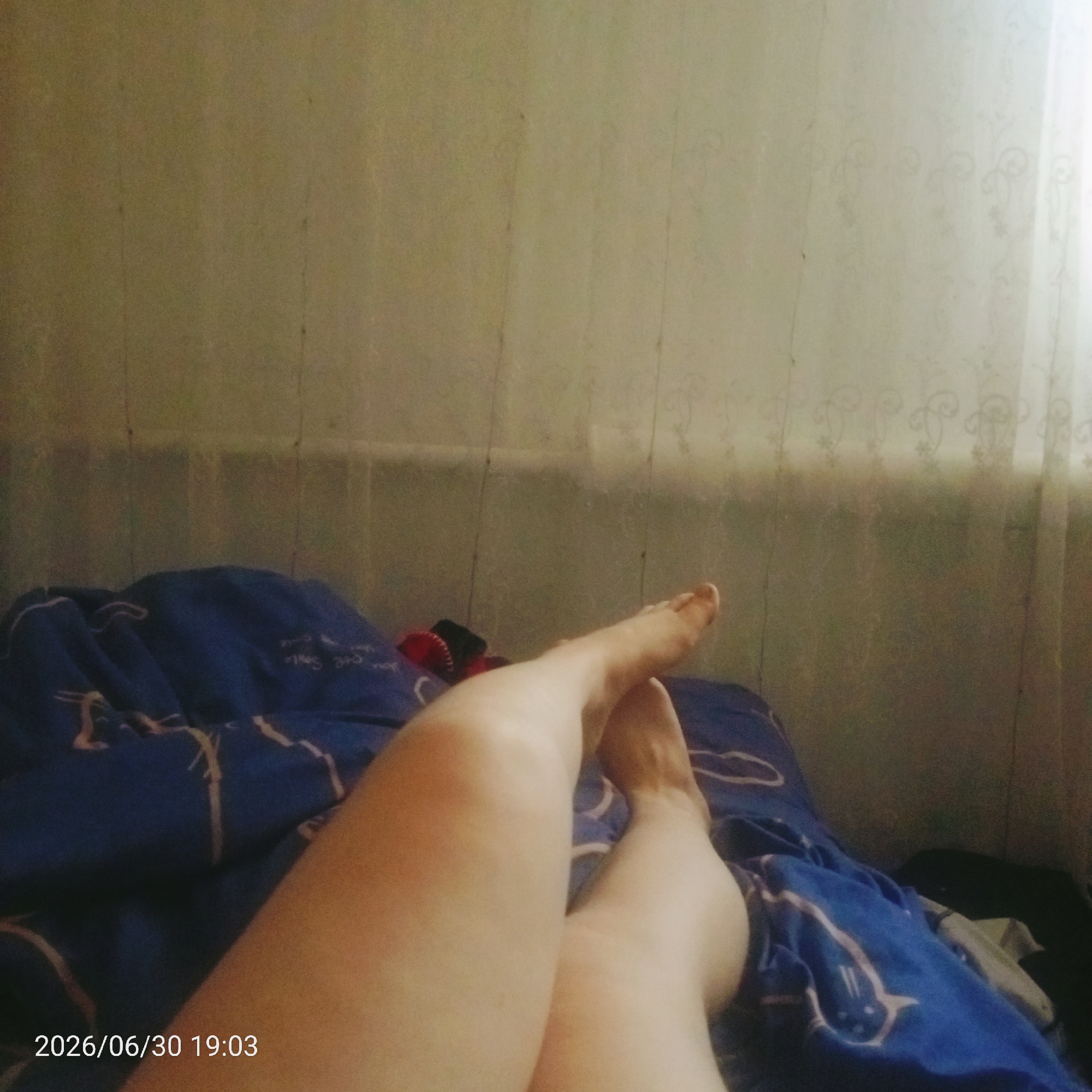 The media file shows a close-up view of a person's legs and feet resting on a blue comforter. The comforter has a pattern of white outlines, possibly depicting stylized animals or figures. The legs are smooth and appear to be bare. The scene is dimly lit, suggesting it might be nighttime or the room is not well-lit. The background is out of focus but appears to be a wall with vertical paneling and a sheer curtain with a subtle pattern. A date stamp in the bottom left corner reads "2026/06/30 19:03," indicating the time the image was captured. The overall impression is a casual and relaxed moment, possibly at home in a bedroom.