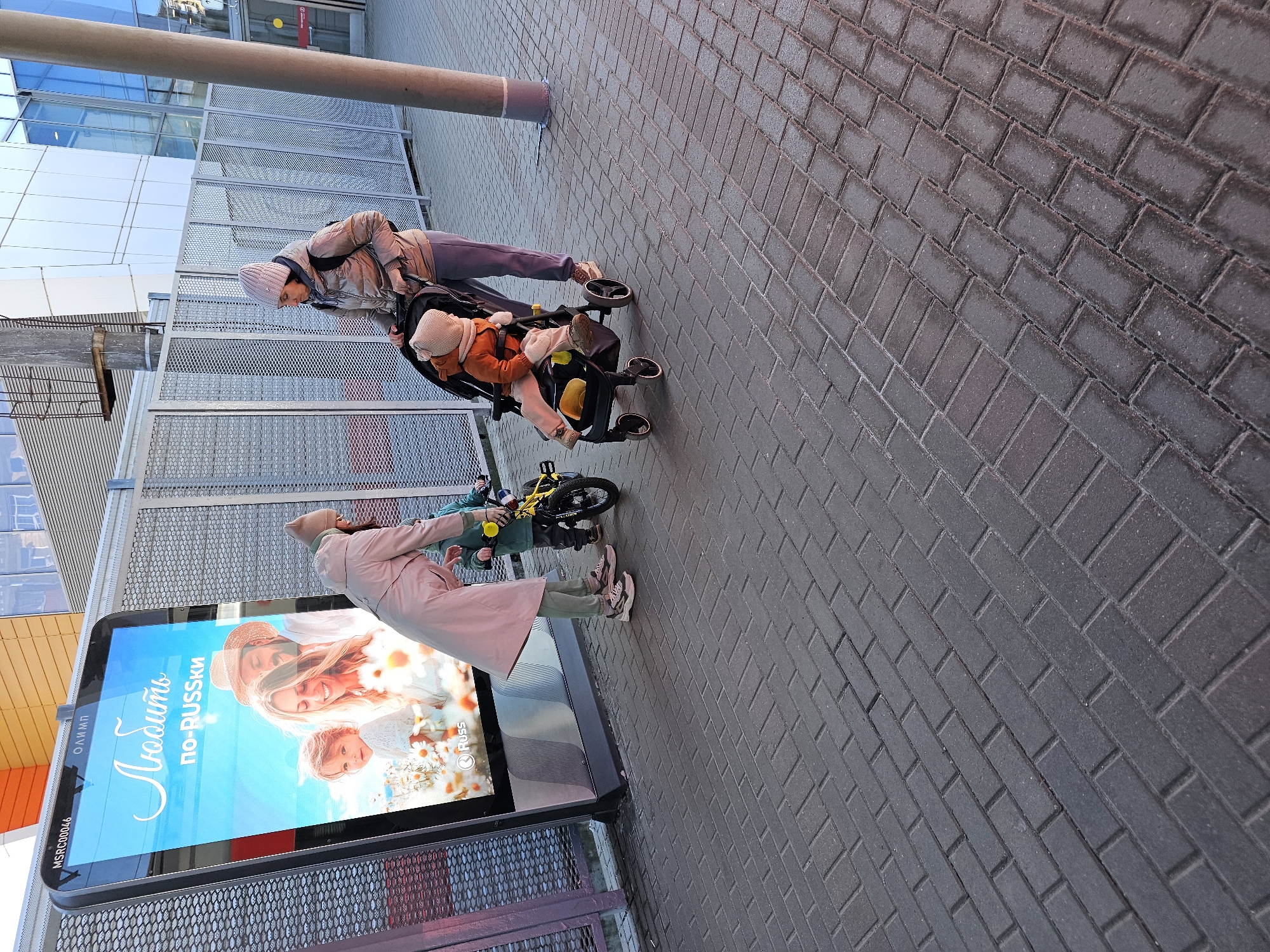 A scene captured in Khimki, Russia, showcases several individuals. In the background, one person is pushing a stroller with a child inside. Further ahead, near a digital advertising screen, another person is tending to a child on a yellow bicycle. The environment consists of a paved brick walkway beside a building with metal grates. The advertising screen displays an image of a fair-haired woman with children.