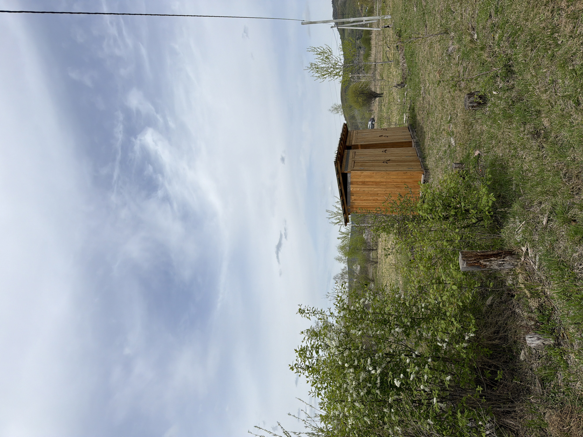 The image shows a small, wooden outhouse with a corrugated metal roof, situated in a grassy area with trees and shrubs. The sky is cloudy. The location is in a rural or semi-rural area, potentially near the town of Теплоозёрск, Russia. There is no evidence of people in the scene. A single car is parked in the background.