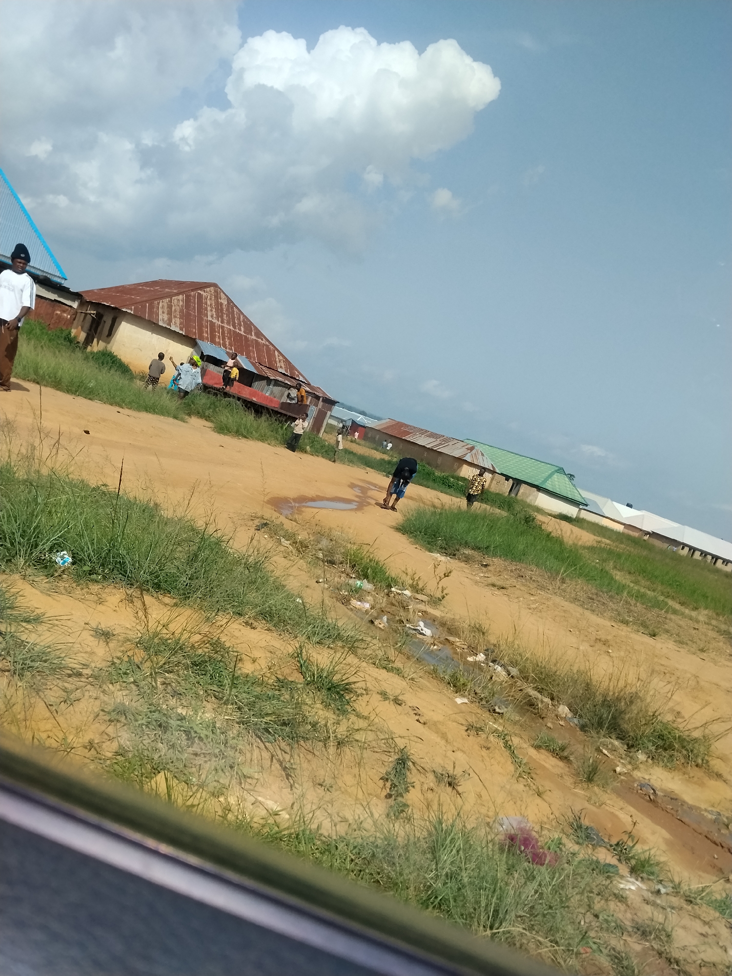The image depicts an outdoor scene in Durumi, Nigeria, characterized by a dirt road with patches of grass and several buildings. The sky is partly cloudy with large cumulus formations. On the left, a person in a white shirt and brown pants stands near a building with a corrugated metal roof, partially obscured by a blue roof. Several other individuals are gathered near this building. Further down the road, another building with a green corrugated metal roof is visible, and a few people are present near it. The road appears to be unpaved and has areas of standing water and scattered debris, suggesting a rural or semi-developed area. The perspective is from a slightly elevated angle, possibly from a vehicle.
