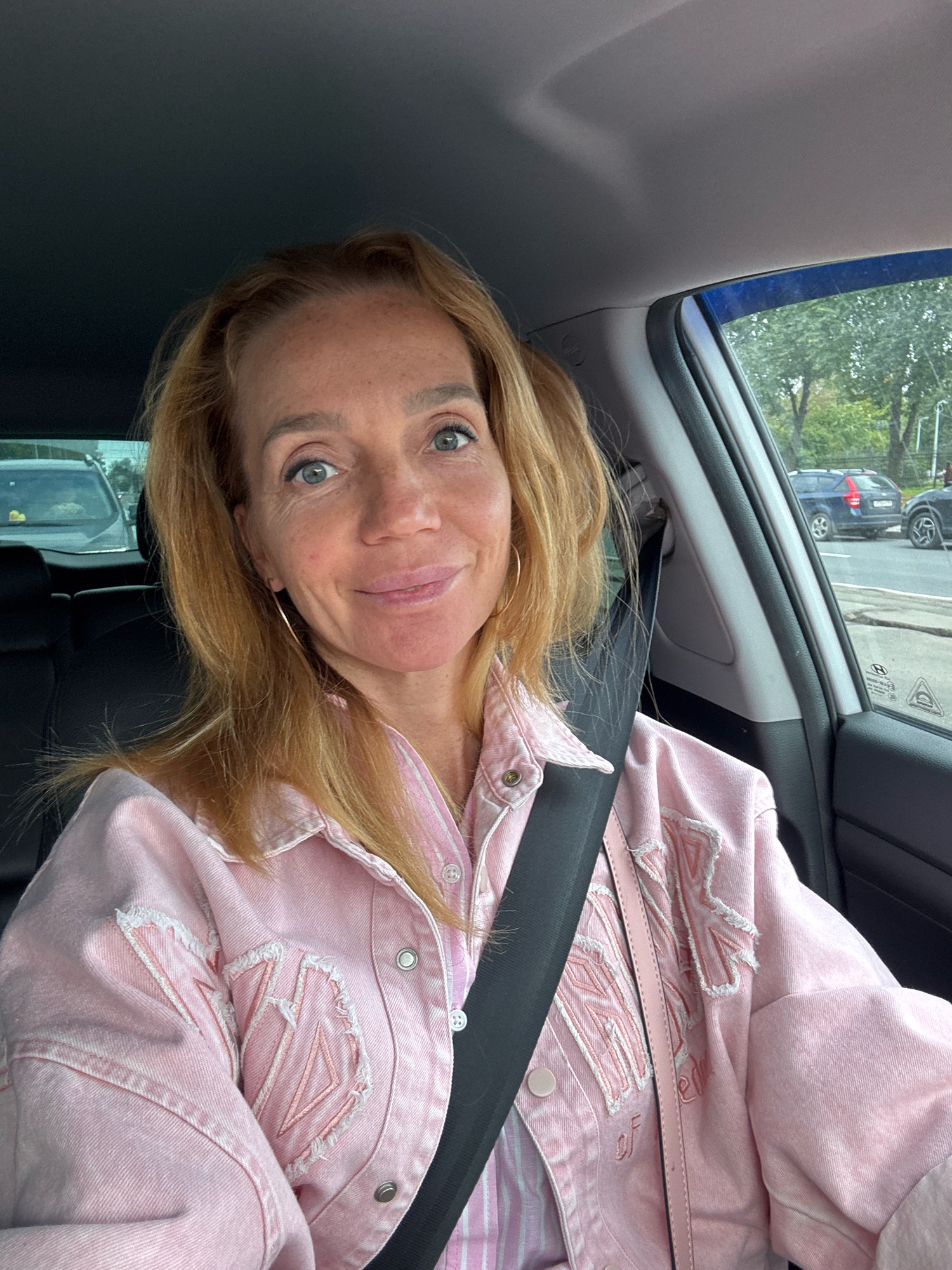 A middle-aged woman with blonde hair and blue eyes is seated in the driver's seat of a car, taking a selfie. She is wearing a pink denim jacket over a striped pink shirt, and her seatbelt is fastened. The interior of the car is visible, including the dashboard and the passenger seat. Outside the car, trees and other vehicles can be seen, suggesting an urban or suburban environment. The woman appears to be smiling and looking directly at the camera. The setting is not identifiable as Saint Petersburg, Russia.