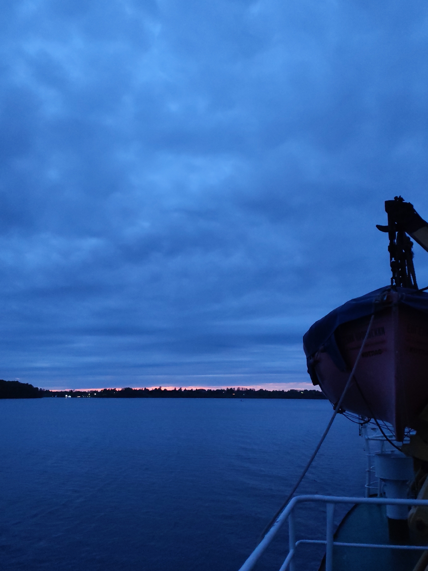 A low-angle shot taken at dusk from the deck of a boat, possibly a ferry, shows a calm body of water, likely a river or a lake. The sky is heavily overcast with a deep blue color, hinting at the fading light of the day. In the distance, a thin strip of land is visible, punctuated with scattered lights, suggesting a shoreline with some habitation. The colors of the horizon indicate the afterglow of sunset or sunrise. To the right side of the frame, a life boat is suspended on a davit system, partially obscuring the view. The details of the life boat are blurred due to the dim light and focus on the distant horizon. The deck railing of the boat and some of its structures are visible in the lower right corner. There are no people visible in the frame. The location cannot be confirmed as Priluki, Russia based on the visual information provided.