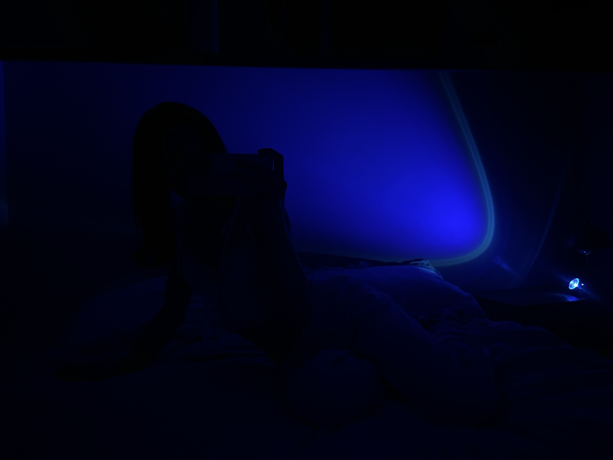 A silhouette of a person is lying on a bed in a dark room, illuminated by a blue light projector. The person is holding a smartphone in their hand, appearing to be looking at it. The projector casts a large, circular blue light pattern on the ceiling or wall, with a green ring around the edge. Another smaller blue light source is visible in the background. The room is sparsely furnished, with the bed covered in white bedding being the most prominent object. The scene suggests a private, indoor setting. No specific landmarks of Ternopil, Ukraine are visible in the image.