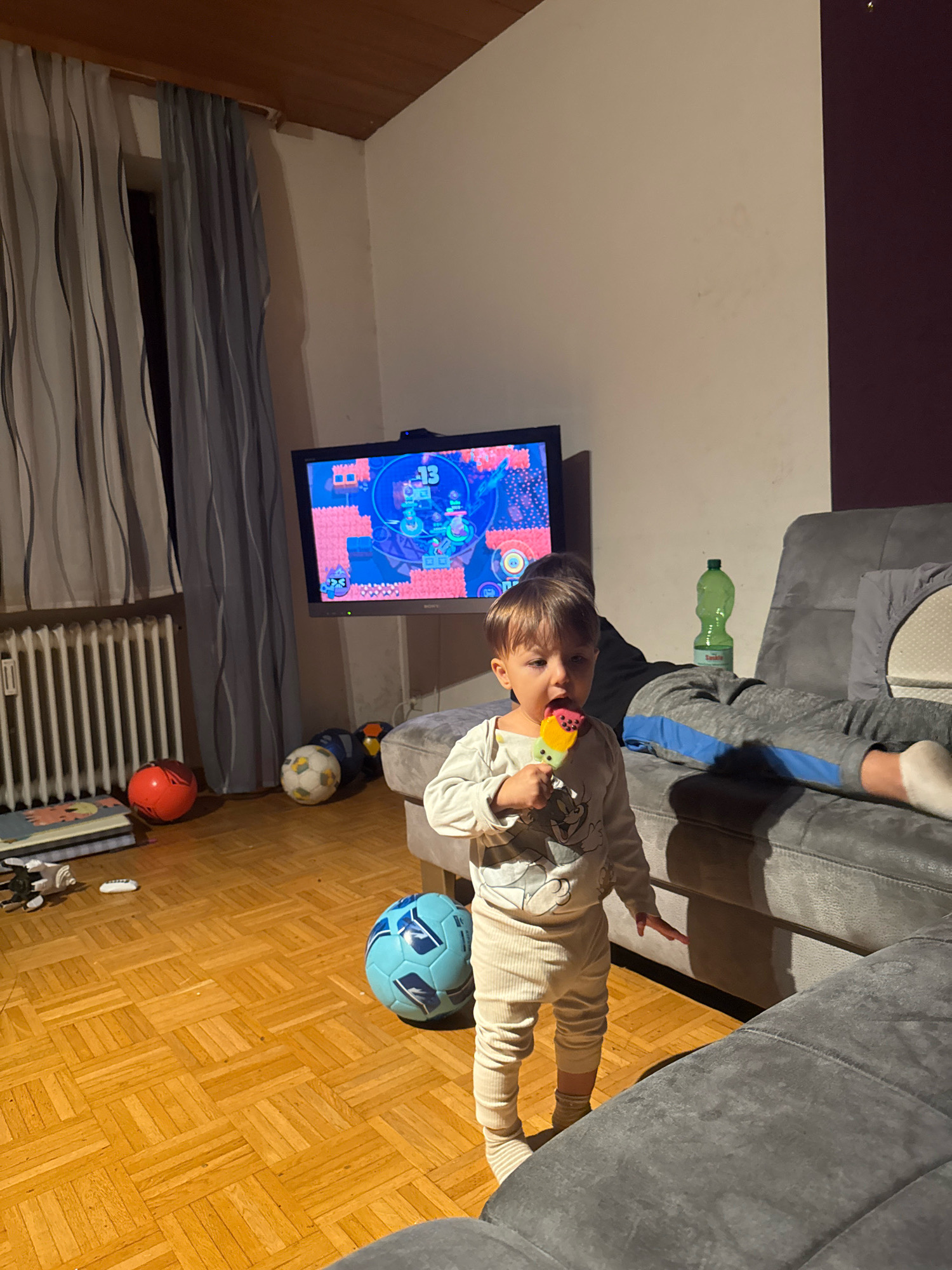 The image depicts an indoor scene, likely a living room, with a young child standing in the foreground holding a colorful popsicle. The child is wearing a light-colored long-sleeved shirt with a cartoon character and ribbed pants with striped socks. The child is looking towards the viewer. Behind the child, a large gray sectional sofa is visible, with another person, possibly an older child or adult, lying down on it wearing gray sweatpants with a blue stripe and white socks. A green bottle of liquid is on the floor next to the sofa.
A television screen displays a video game with colorful graphics. In the background, to the left, a radiator is visible beneath a window covered by vertical striped curtains. Several balls are scattered on the wooden floor, including a red ball, a white and black soccer ball, and a blue soccer ball. A stack of books or mats is near the radiator. The overall lighting suggests it is evening or a dimly lit room. The scene does not provide specific geographical markers for the city of Überherrn, Germany.
