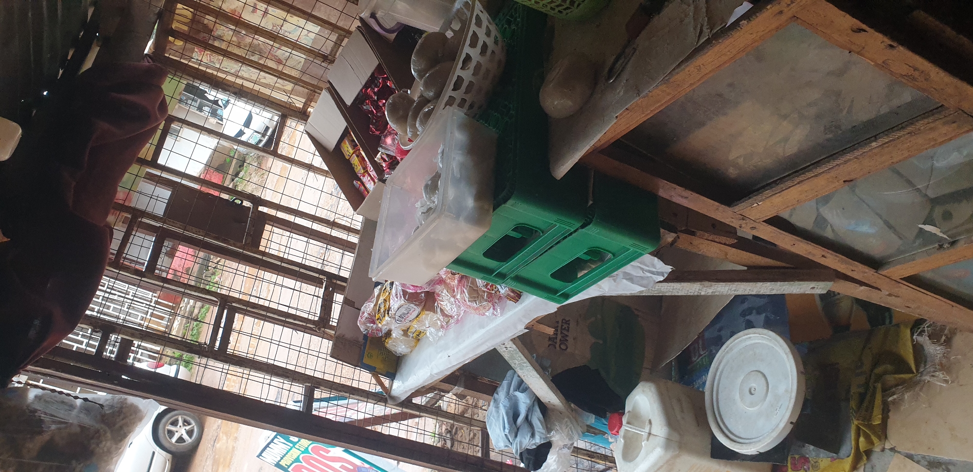 The image depicts the interior of a small, cluttered kiosk or open-air stall, likely a provision store, in Sabon Tasha, Nigeria. The structure appears to be made of salvaged materials, with a corrugated metal roof and a cage-like metal bar front. A table, covered with a white plastic sheet, displays various goods. These include packaged snacks or biscuits in clear plastic bags, some loose items in a clear plastic container, and a basket filled with round objects, possibly fruits or vegetables. Two green plastic crates are stacked on the table, one holding a clear plastic container with some white, crumpled items. Behind the table, several cardboard boxes are stacked, some containing packaged goods with red and yellow wrappers. To the left of the table, a white plastic water dispenser with a red lid sits on the ground, next to a grey circular lid and a pile of fabric. A large cardboard box with text reading "POWER" is visible leaning against the wall behind the table. The overall impression is one of a busy, perhaps somewhat disorganized, retail space. The background suggests an outdoor environment with a road and buildings visible through the metal bars.