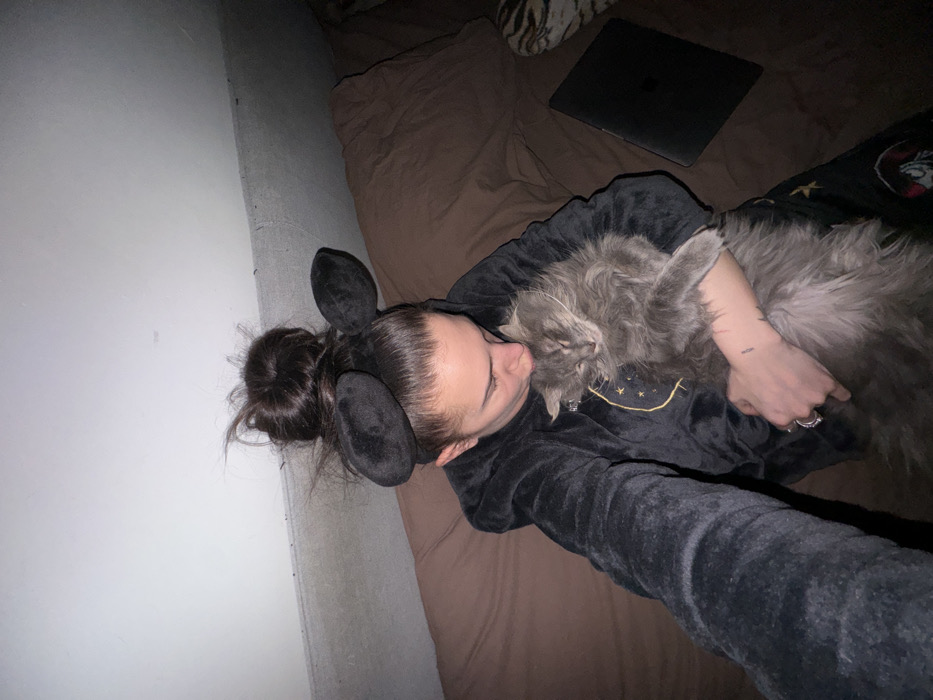 The image captures a cozy and intimate moment between a person and a cat in what appears to be a dimly lit indoor setting, likely a bedroom.
A person, seemingly a young woman, is lying on her back on a bed with brown or dark-colored bedding. She is wearing a dark, possibly plush or fleece, hooded garment that features two rounded, dark, ear-like attachments on the hood, resembling animal ears. Her dark hair is styled in a messy bun. Her face is visible in profile, turned towards the cat, with her eyes appearing to be closed or looking down. She has a faint, small tattoo on her left forearm and is wearing several rings on her left hand. Her left arm is wrapped around the cat.
Resting on the person's chest and stomach is a large, very fluffy, grey cat. Its head is nuzzling or rubbing against the person's face, with its eyes appearing closed, suggesting comfort and affection. The cat's long, shaggy fur is prominent.
The activity depicted is a moment of affectionate cuddling between the person and their cat, conveying a sense of warmth and companionship.
The background shows a light-colored, possibly white or off-white, wall on the left side. Behind the person's head, on the bed, a dark-colored laptop is visible. A patterned textile, possibly a pillow or blanket with an animal print, is partially visible in the upper right corner. The lighting in the image is dim, suggesting it might be evening or nighttime. On the person's dark top, there is a subtle yellow embroidered design resembling a simple smiley face (a curved line with two dots above it). No discernible text is visible in the image.