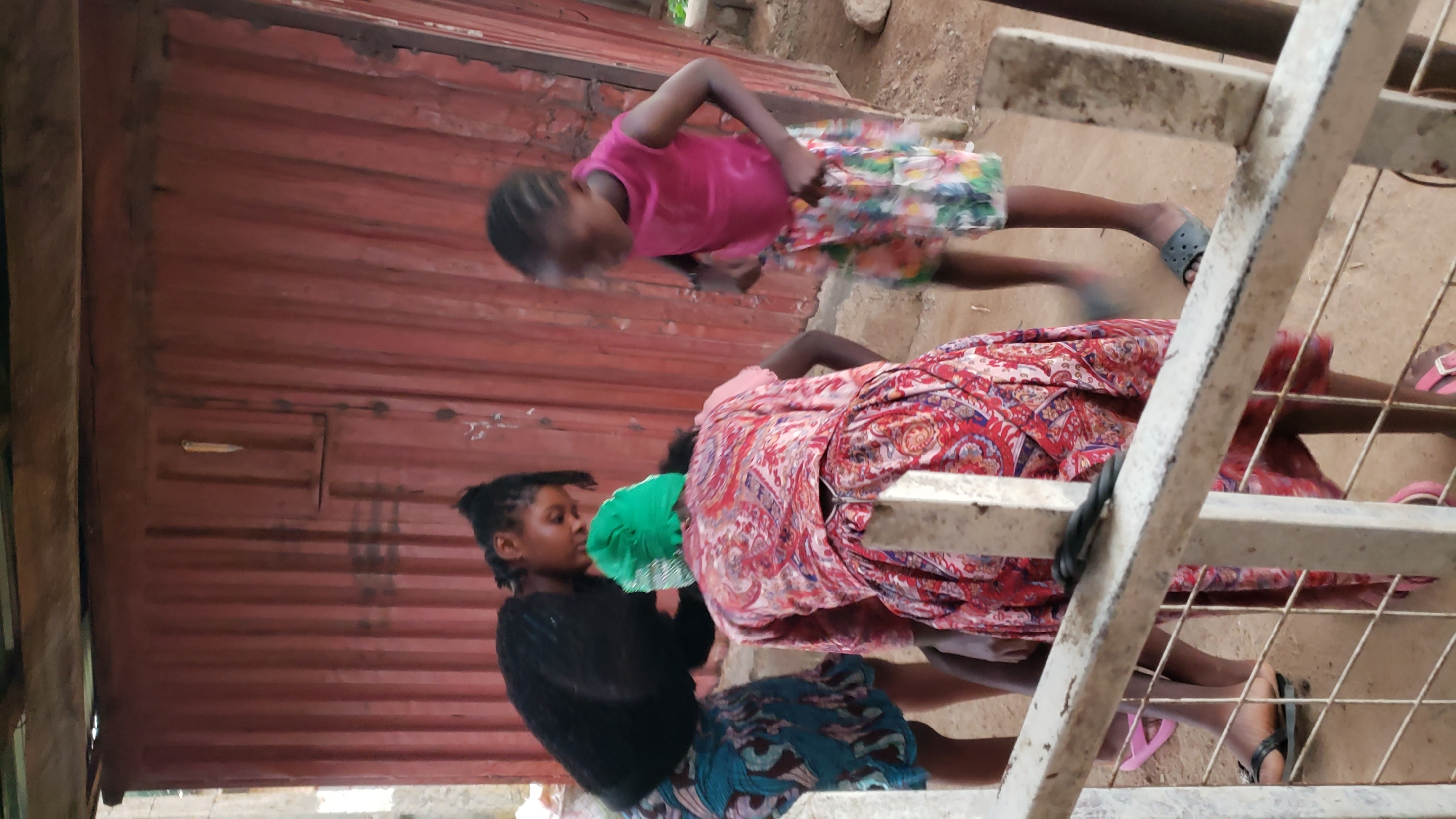 In Narayi, Nigeria, three individuals are visible outdoors, positioned near a corrugated metal structure. The person in the foreground, wearing a red and white patterned dress and a green headscarf, is bent over. To their left, a person in a black top and a blue and brown patterned skirt is also bent forward, appearing to interact with the individual in the red dress. To the right, a younger person in a pink sleeveless top and a colorful patterned skirt stands, looking down. A white metal fence with a diamond-shaped mesh separates the viewer from the scene. The ground is dirt.