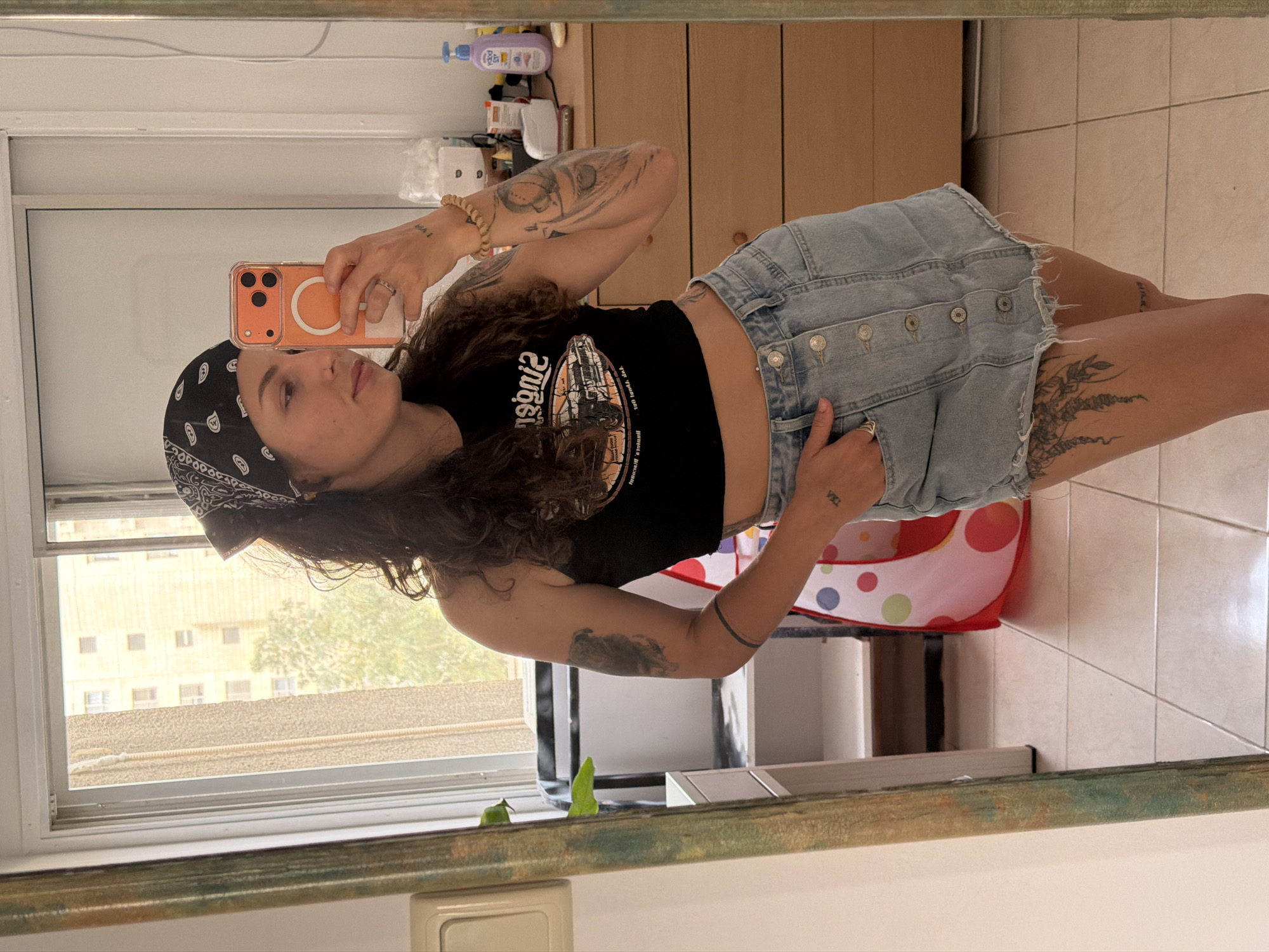 A woman stands indoors, reflected in a mirror. She wears a black crop top, a denim skirt, and a black bandana. Her left arm is raised, holding a phone with an orange case, presumably taking the photograph. Tattoos are visible on her arms and legs. Behind her, a dresser with wooden drawers is partially visible, along with assorted items on top of it. A window shows a view of a building across the street, likely in an urban setting. Part of a children's toy is also visible on the floor. The scene suggests a domestic interior. While the location is claimed to be Kiryat Bialik, Occupied Palestine, this cannot be verified from the image.