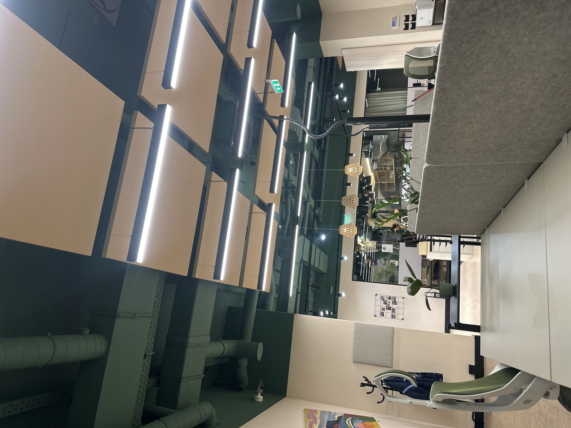 The image depicts a modern office or co-working space located in Budva, Montenegro. The scene is well-lit and features a contemporary design with exposed industrial elements and acoustic treatments.
The ceiling displays an industrial aesthetic with dark green exposed HVAC ducts, pipes, and electrical conduits. Interspersed among these are numerous light-colored, rectangular acoustic panels and multiple linear LED light fixtures providing illumination. A green "EXIT" sign with a white arrow and figure is visible in the upper mid-right. A white security camera (CCTV) is mounted on the ceiling in the upper-left quadrant. In the background, three woven wooden pendant light fixtures hang over an adjacent area.
The walls are painted a light, neutral beige. On the left wall, a colorful abstract painting is displayed above a black coat rack holding a dark blue jacket. Adjacent to the coat rack, a grey rectangular sound-absorbing panel is mounted, and further right, a wire grid displays multiple small photographs.
The foreground is dominated by a white desk surface, with a light green and white ergonomic office chair positioned to its left. Grey felt desk dividers create a cubicle-like arrangement. Behind these dividers, a tall black pole with a flexible, segmented arm extends upwards.
In the mid-ground, a black table holds a green potted plant. Other smaller green potted plants are visible behind the desk dividers. Through a large glass partition in the background, another section of the office is visible, featuring additional desks, chairs, and shelving units. A white printer or scanner is discernible on a shelf in the background right. A blurry, partial human figure is faintly visible in the deeper background through the glass partition. The flooring appears to be light-colored tile.