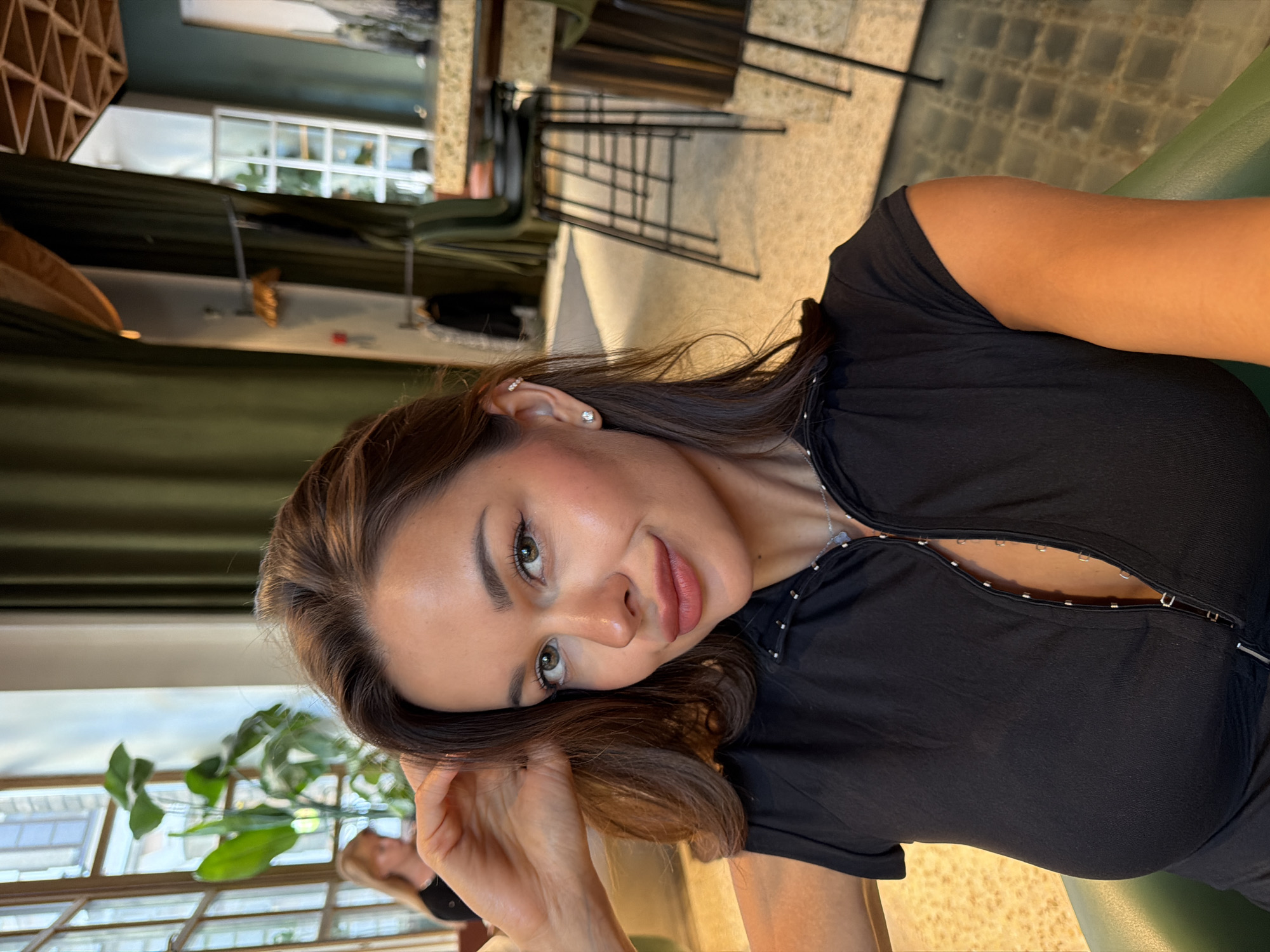 The image displays a close-up, front-facing portrait of an adult female, likely a selfie, taken in an interior setting in Moscow, Russia. The woman, appearing to be in her 20s or 30s, has medium brown hair styled with loose waves and parts slightly to her right. She has light green eyes, arched eyebrows, and prominent cheekbones. Her skin is tanned, and she wears subtle makeup, including a rosy blush, eyeliner, and pinkish-brown lip color. Her left hand is raised, with fingers lightly touching her hair near her right temple.
She is wearing a short-sleeved black top with a distinctive front closure featuring multiple small, silver hook-and-eye clasps running vertically down the center from the collar, revealing a hint of cleavage. A delicate silver necklace with a small, light-colored pendant hangs around her neck. In her visible left ear, she wears a small silver stud earring in the lobe and a silver ear cuff with small stones positioned higher on the helix.
The background depicts a stylish indoor space, suggestive of a cafe, restaurant, or lounge. To the left, large multi-pane windows with arched tops allow natural light into the room, revealing a large green leafy plant, possibly a Monstera, positioned near the window. A blurred figure of another woman with blonde hair is seated in the far left background. To the right, heavy dark green curtains drape along a wall. The room is furnished with high tables and dark green upholstered bar stools featuring black metal legs. The flooring is a light beige or off-white speckled terrazzo or aggregate material. Wall-mounted light fixtures with metallic accents are also visible in the background.