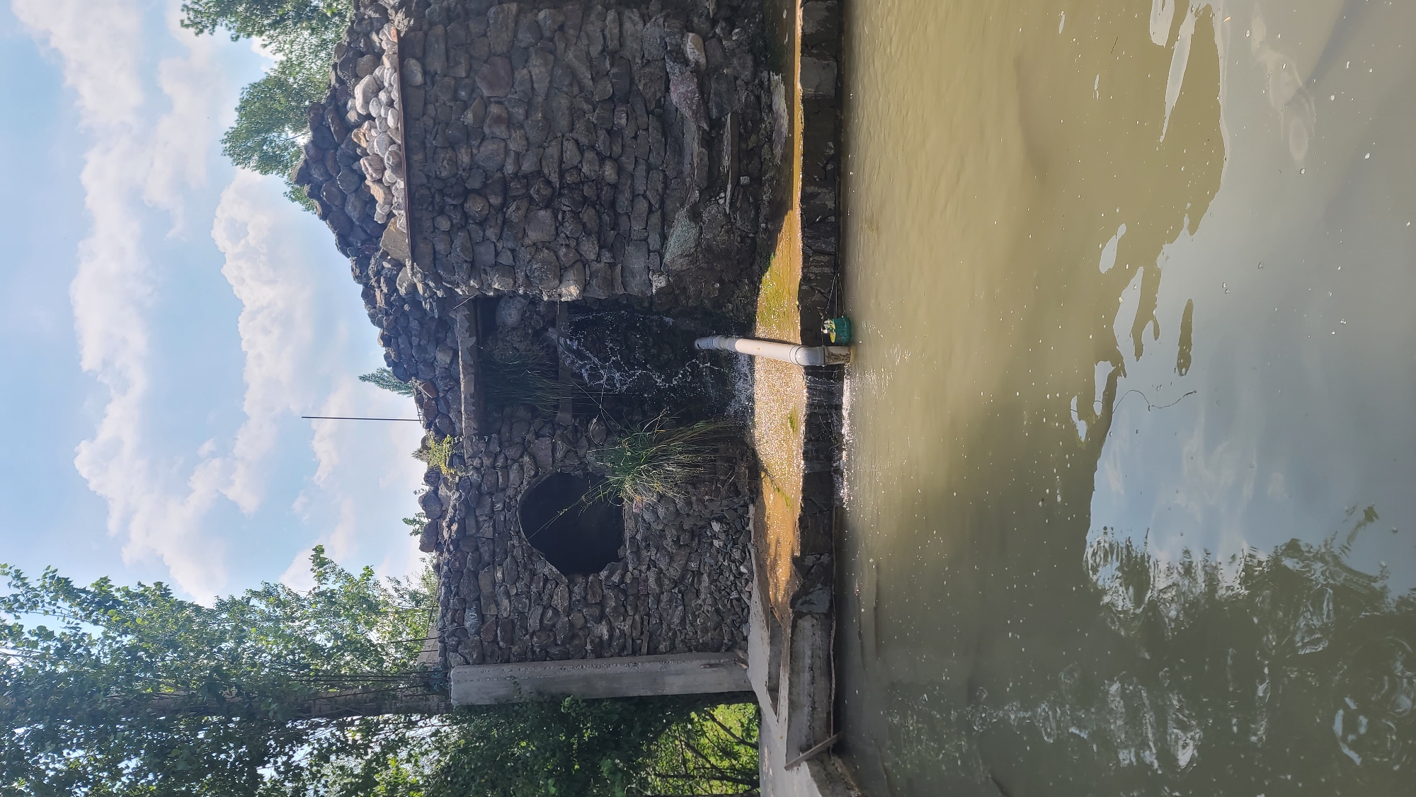 A stone structure with a small waterfall is depicted in Arstanbap, Kyrgyzstan. The structure appears partially ruined, with rough-hewn stones forming its walls and roof. A pipe protrudes from the waterfall, directing water into a murky pool in the foreground. Green foliage surrounds the structure, and a clear sky with scattered clouds is visible above. The scene appears to be an outdoor area, possibly a park or a natural attraction.