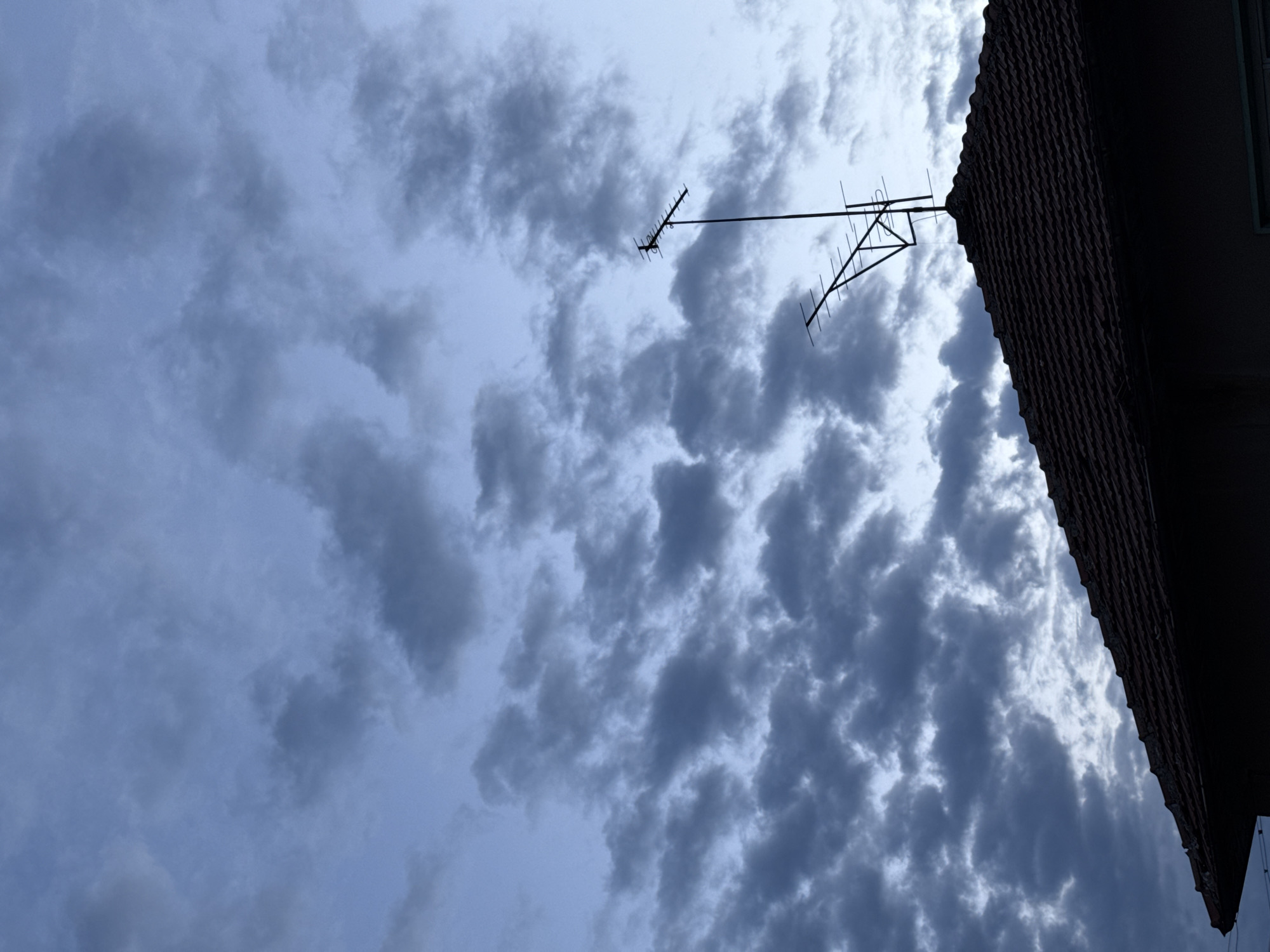 The image shows a view looking up at the sky, filled with grey and white clouds. A rooftop with dark red tiles is visible in the upper right corner of the frame. A television antenna is mounted on the rooftop, reaching into the cloudy sky. The location is Pleven, Bulgaria.
