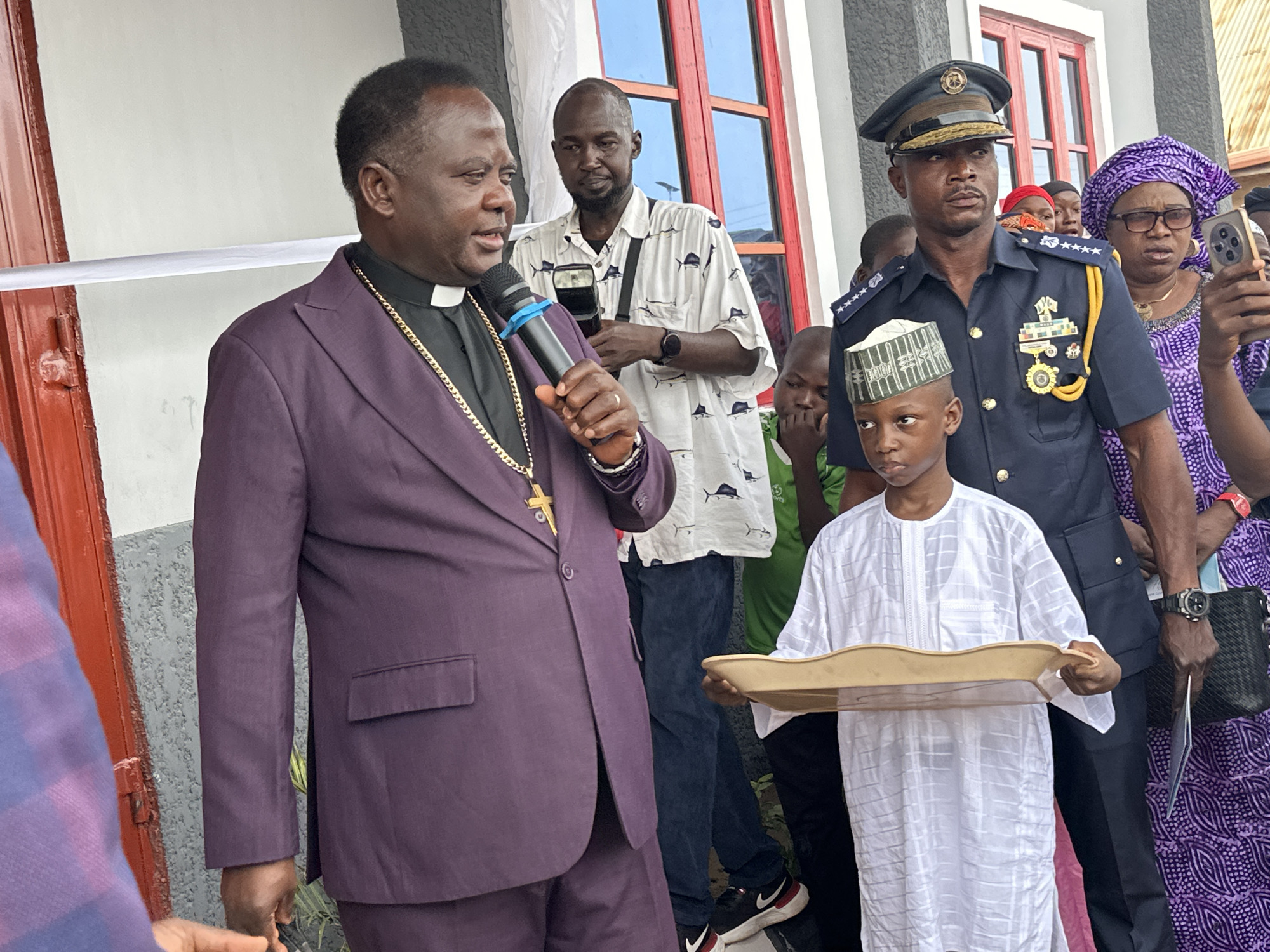 A man in a purple suit with a clerical collar and gold cross necklace speaks into a microphone in front of a building in Goni Gara, Nigeria. A man in a white shirt with black fish designs stands behind him holding a camera. A boy wearing a white patterned tunic and a white skull cap holds a tan tray. To the right of the boy, a man in a dark blue uniform with gold buttons and insignia wears a peaked cap. Behind him, a woman in a purple patterned dress and headwrap holds a cell phone. A white ribbon stretches across the door of the building behind the man in the purple suit. The building has gray stucco walls and red door and window frames.