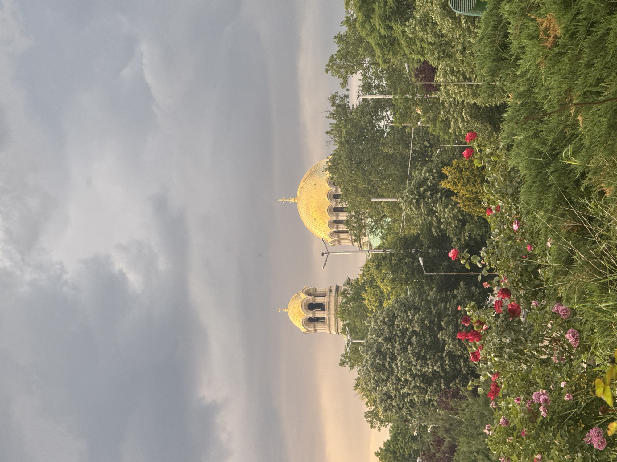 An outdoor scene captures a grand architectural structure, likely a cathedral, set against a dramatic, cloudy sky and surrounded by lush greenery. Two prominent golden domes, characteristic of Orthodox Christian architecture, dominate the left side of the frame. The larger dome is a central, onion-shaped structure, glistening with a golden hue, suggesting it's illuminated by a warm light, possibly from a breaking sunbeam. To its right, a smaller golden-domed tower with arched openings is visible above the treeline.
The sky is heavy with dark grey, overcast clouds, indicative of stormy weather or its recent passing. However, a lighter, yellowish glow is present near the horizon line, illuminating the domes. The foreground and right side of the image are filled with diverse green foliage, including tall trees and dense bushes. Vibrant clusters of red and pink/purple roses are prominent among the bushes, adding splashes of color. Partially visible streetlights are interspersed among the trees. A light green slatted object, possibly a bench, can be seen through the trees in the upper right background. No people or visible text are present in the image. The architectural style, combined with the location hint, strongly suggests the Alexander Nevsky Cathedral in Sofia, Bulgaria, possibly photographed during a dramatic sunrise or sunset after a cloudy period.