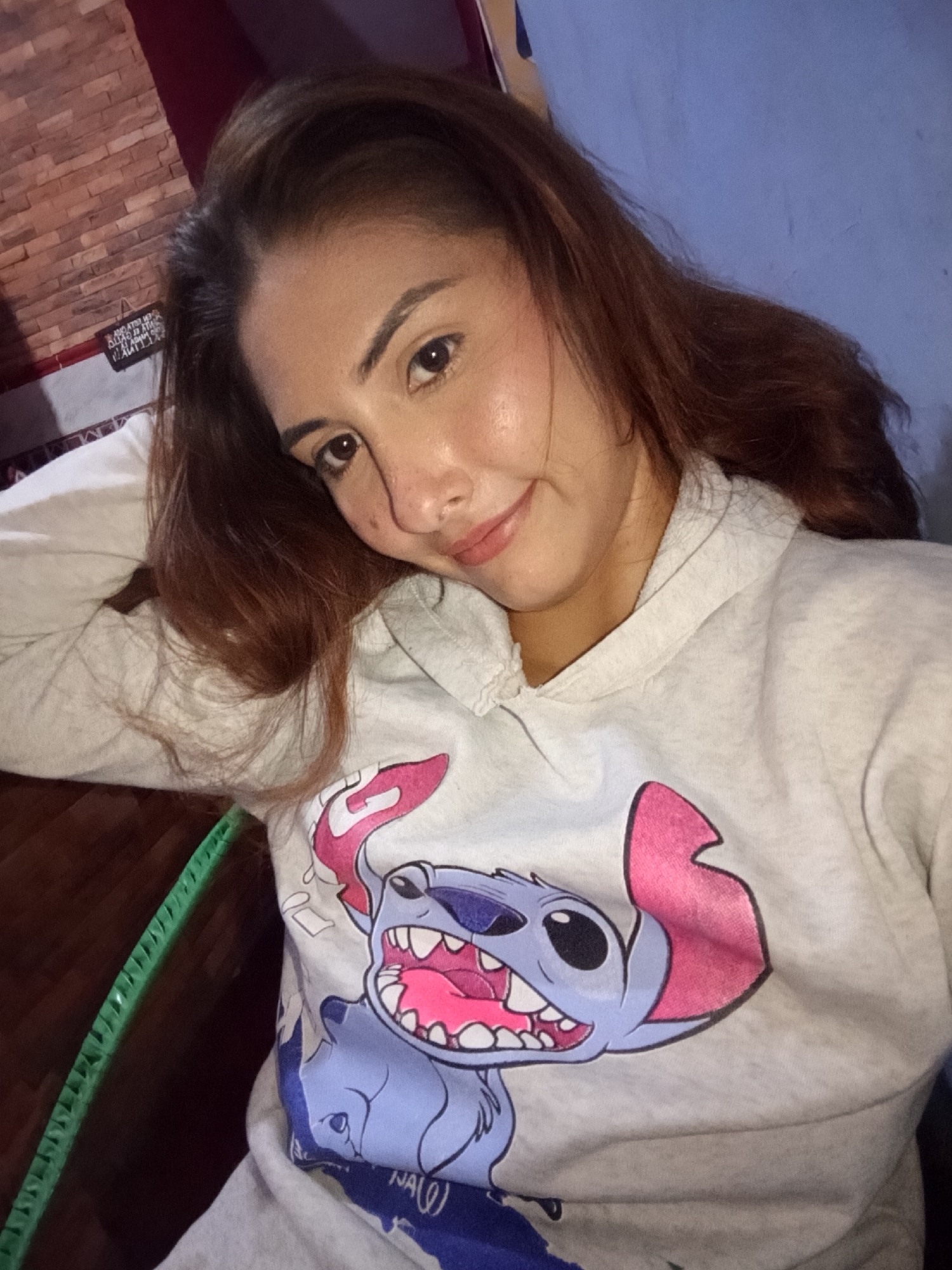 An adult female is depicted in a selfie, looking directly at the camera with a subtle smile. She has medium-long, wavy dark brown hair, dark brown eyes, and light-medium skin tone, with her head tilted slightly to her left. She wears a light grey hooded sweatshirt featuring a large graphic of the Disney character Stitch, with his mouth open, pink tongue visible, and a prominent pink inner ear. Partial blue text, possibly "NA(0)", is visible beneath the character. The background is an indoor setting with two distinct wall sections. To the left, there is a reddish-brown brick-patterned wall. An inverted sign with text, partially legible as Spanish words like "GRATIS," is affixed to this wall. To the right, a solid periwinkle blue wall is visible. In the lower-left corner, a bright green, ridged, flexible object, possibly a hula hoop or similar item, is present, partially obscured by a dark wooden surface. The scene is located in the city of Juan Griego, Venezuela.