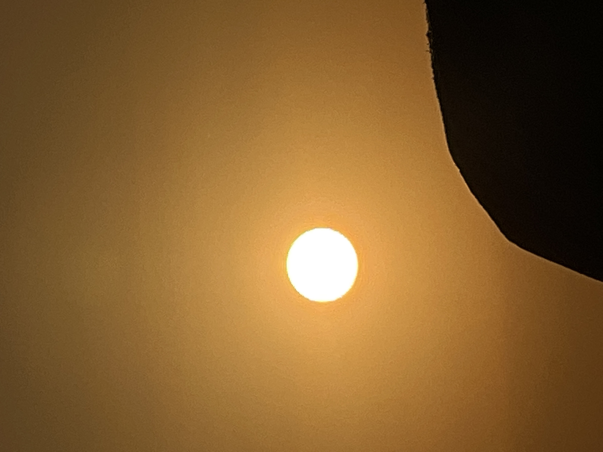 The image shows a bright, circular sun against an orange sky. In the foreground, a dark silhouette of a hill or mountain is visible. The sun is positioned in the upper left quadrant of the image, while the silhouette occupies the lower right portion. The scene appears to be a sunset or sunrise over a landscape, possibly near Kuta, Indonesia, given the warm, hazy atmosphere typical of tropical environments. There are no people or specific objects of note other than the natural elements depicted.
