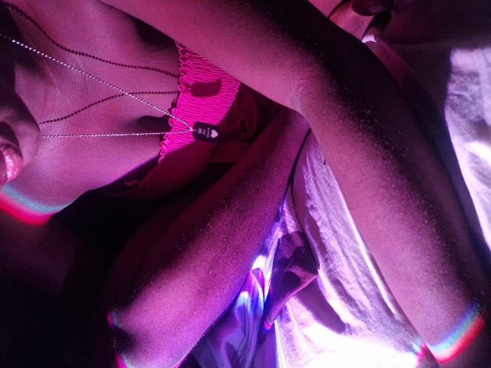 An individual lies down, bathed in pink and purple light. A thin, silver necklace drapes across their chest, bearing a small tag. The person's arms are positioned close to the body. The surface beneath them appears to be a bed with a white sheet. The lighting creates rainbow-like glows at the edges of the person's body. The overall impression is intimate.