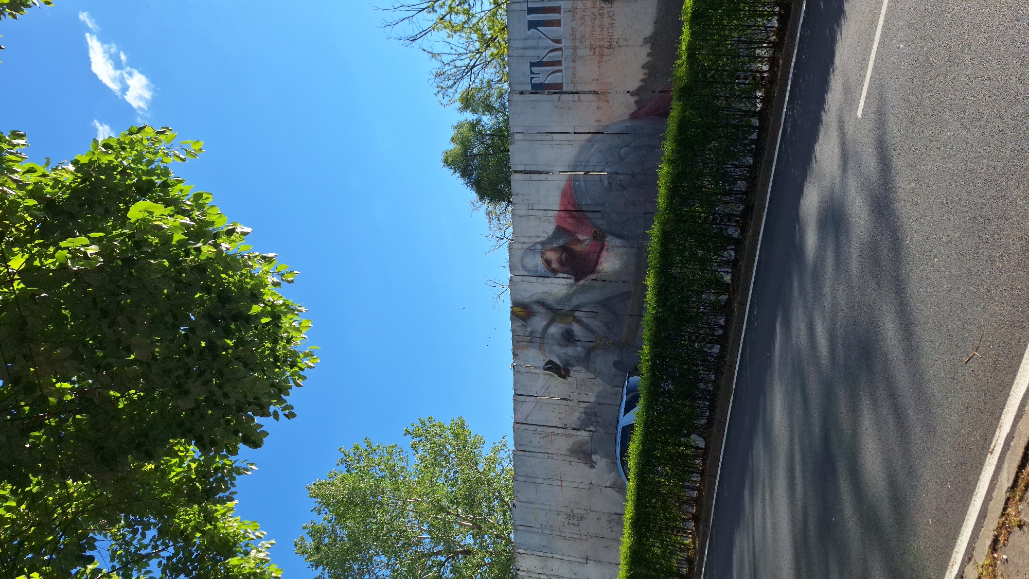 The image shows a section of the Berlin Wall, now located in Blagoevgrad, Bulgaria. The wall is adorned with a mural depicting a medieval knight on horseback. A narrow hedge runs along the base of the wall, separating it from a two-lane road. The sky is blue with scattered clouds. Green trees flank the left side of the image.