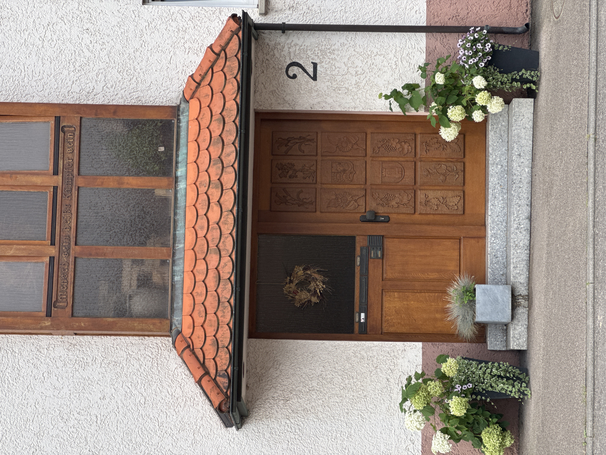 This is a photograph of the front entrance of a house located at number 2, Hauptstraße 2, in Bietigheim-Bissingen, Germany. The entrance features a wooden door with intricate carvings in a grid pattern. Above the door, a small red-tiled awning is present. To the left of the door, a small glass pane is visible, and a circular, dried-flower wreath hangs within it. The house exterior is textured white stucco with a band of pink paint below the awning. The number "2" is prominently displayed in black on the stucco to the right of the door. Two planters are situated on the steps leading to the entrance: one to the left contains large white and green hydrangeas and trailing greenery, and another, smaller grey planter to the right holds ornamental grass. The ground in front of the steps is paved with grey concrete. A wooden frame with multiple glass panes is situated above the awning, containing carved text in German: "Mensch sein heißt Kämpfer sein" which translates to "To be human means to be a fighter". The lighting suggests it is daytime, possibly overcast.
