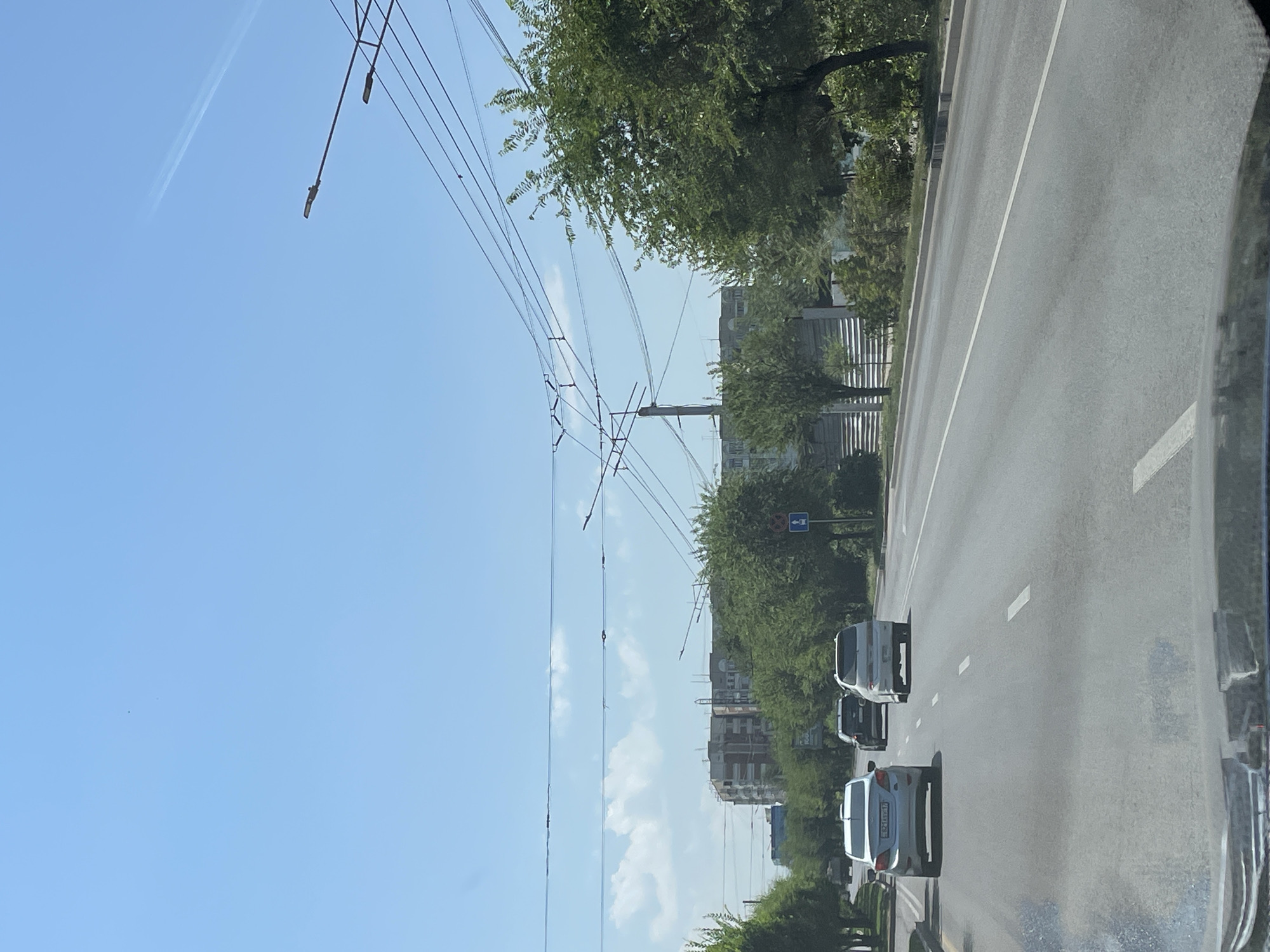 The image shows a view from a vehicle traveling on a multi-lane road in Almaty, Kazakhstan. The sky is clear blue with a few wispy clouds and a faint contrail from an aircraft. Overhead, power lines crisscross the sky, supported by poles. On either side of the road, trees line the street, with buildings visible in the background. Several cars are driving on the road, including a white Hyundai Solaris in the foreground, a black car behind it, and another white car further ahead. A blue traffic sign with a white arrow is visible on the right side of the road. The overall scene depicts a typical urban environment on a sunny day.
